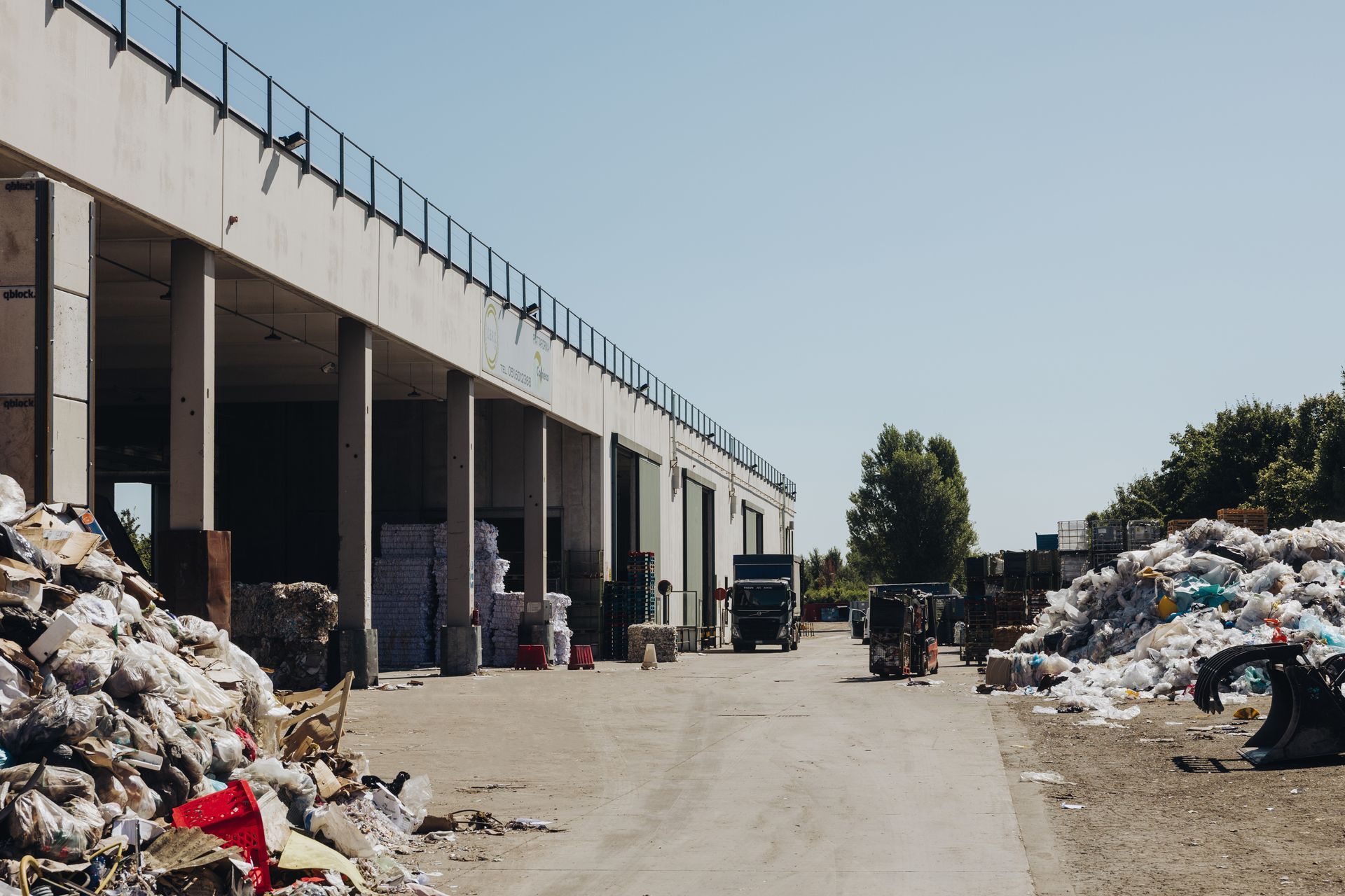 Area industriale con cumuli di materiali riciclabili selezionati accanto a un lungo edificio adibito a magazzino.