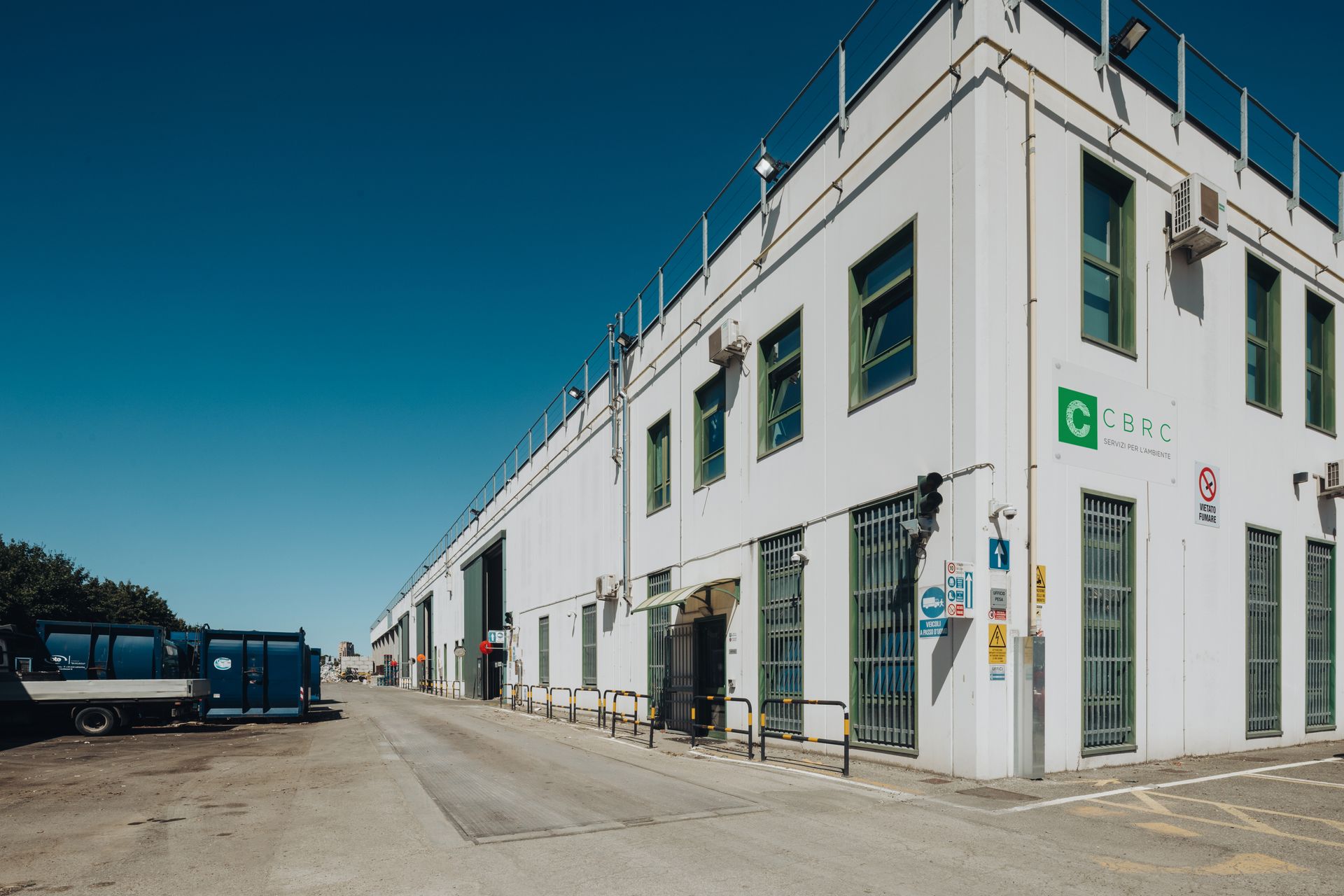 Edificio industriale bianco con insegne verdi su una strada soleggiata, con camion parcheggiati a lato.