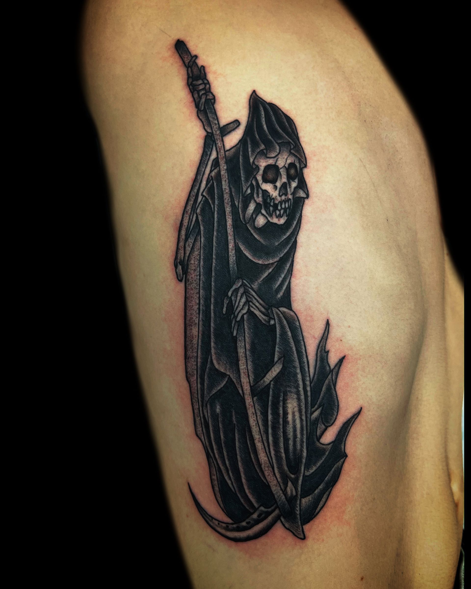 Grim Reaper tattoo on torso, holding a scythe. Black ink with detail, on fair skin.