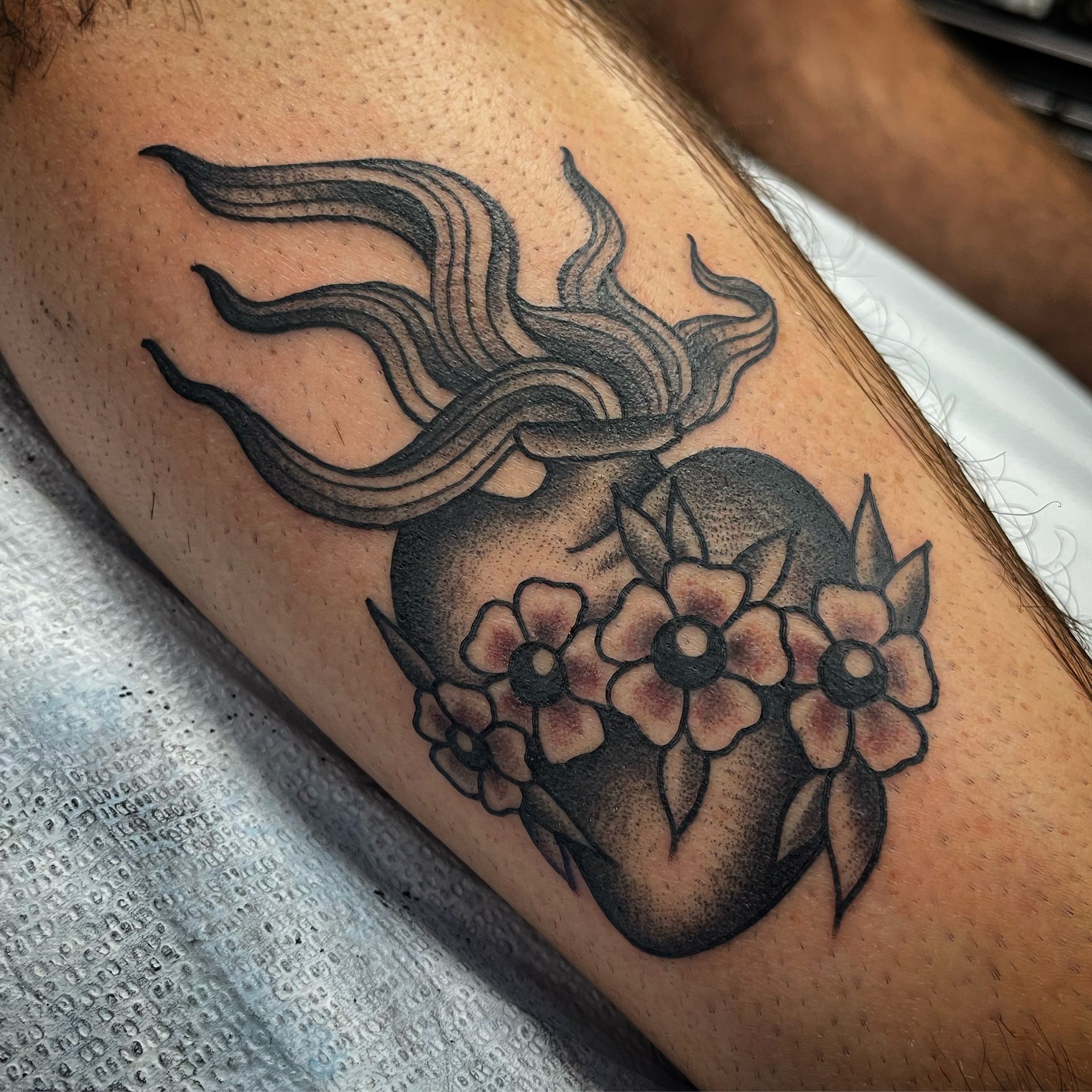 Tattoo of a black heart with flowers and flames on a person's leg, in black and gray.
