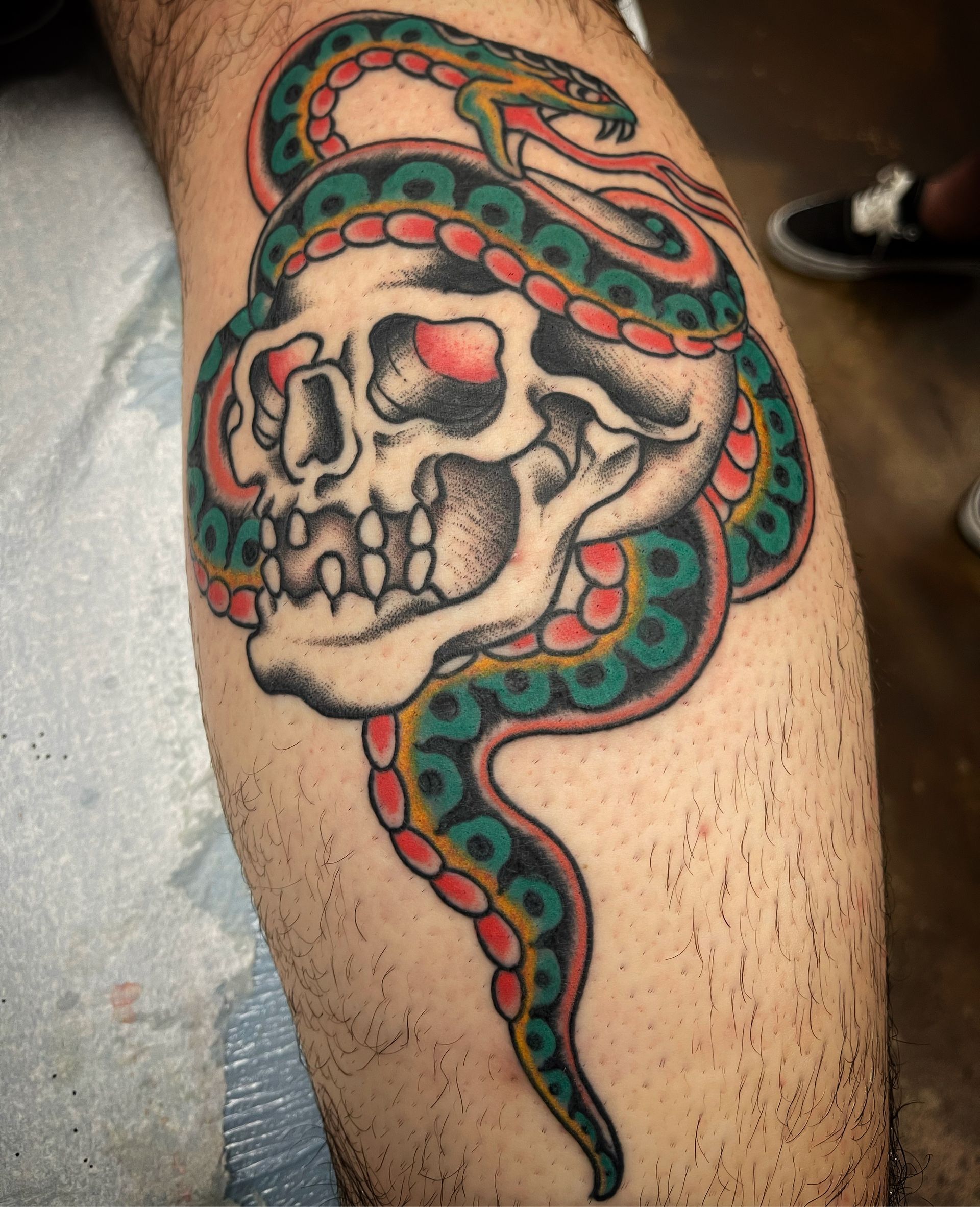 Tattoo on calf: Skull wrapped by a green snake, with red details.
