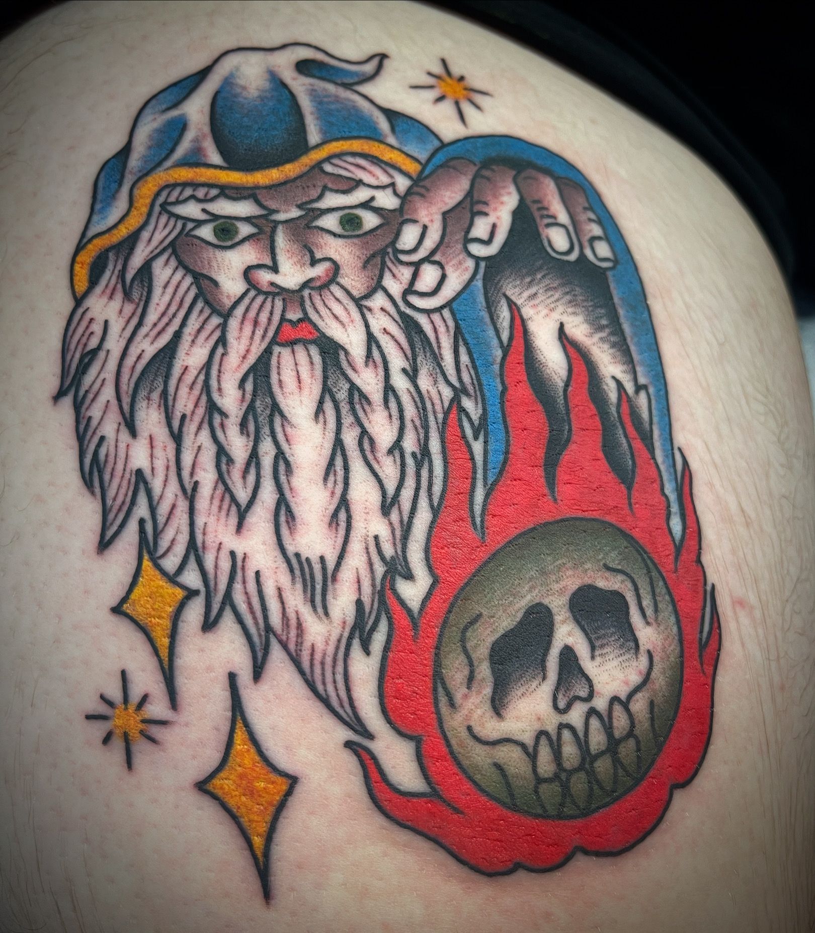 Wizard tattoo, holding skull engulfed in red flames, with blue hat, surrounded by yellow stars.