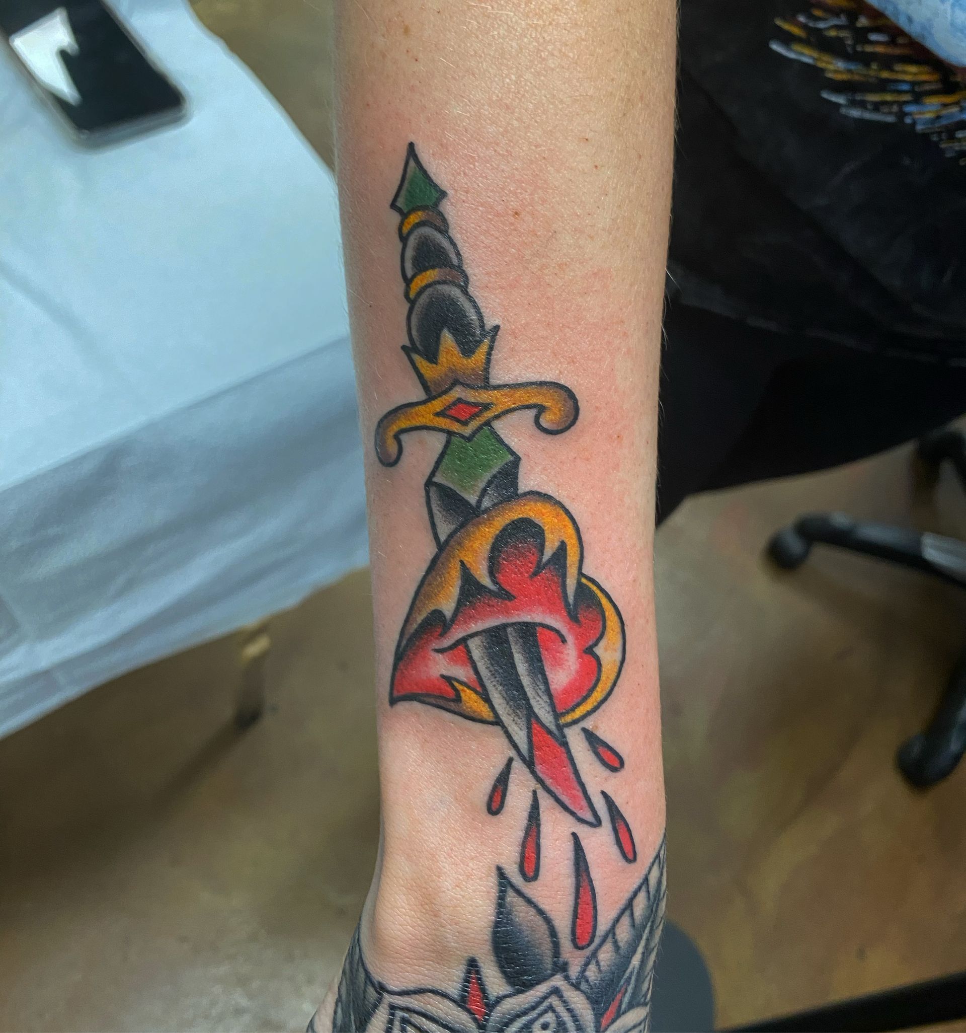 Tattoo of a dagger piercing a red heart with blood dripping. Black, yellow, green details.
