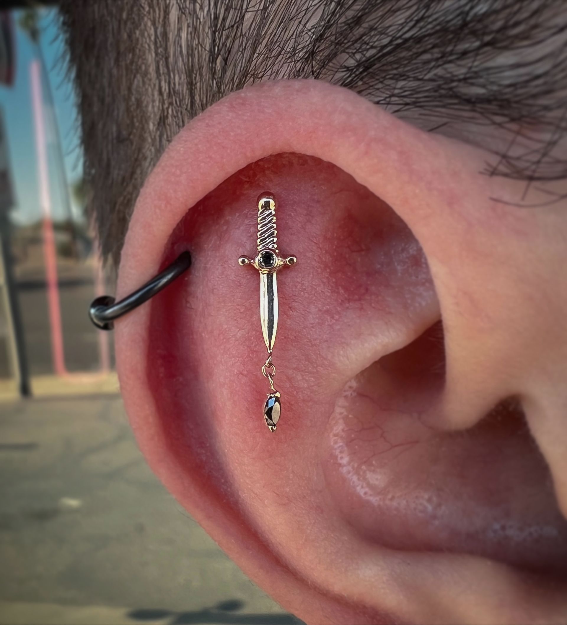 Close-up of an ear with a silver sword-shaped helix piercing and black hoop jewelry