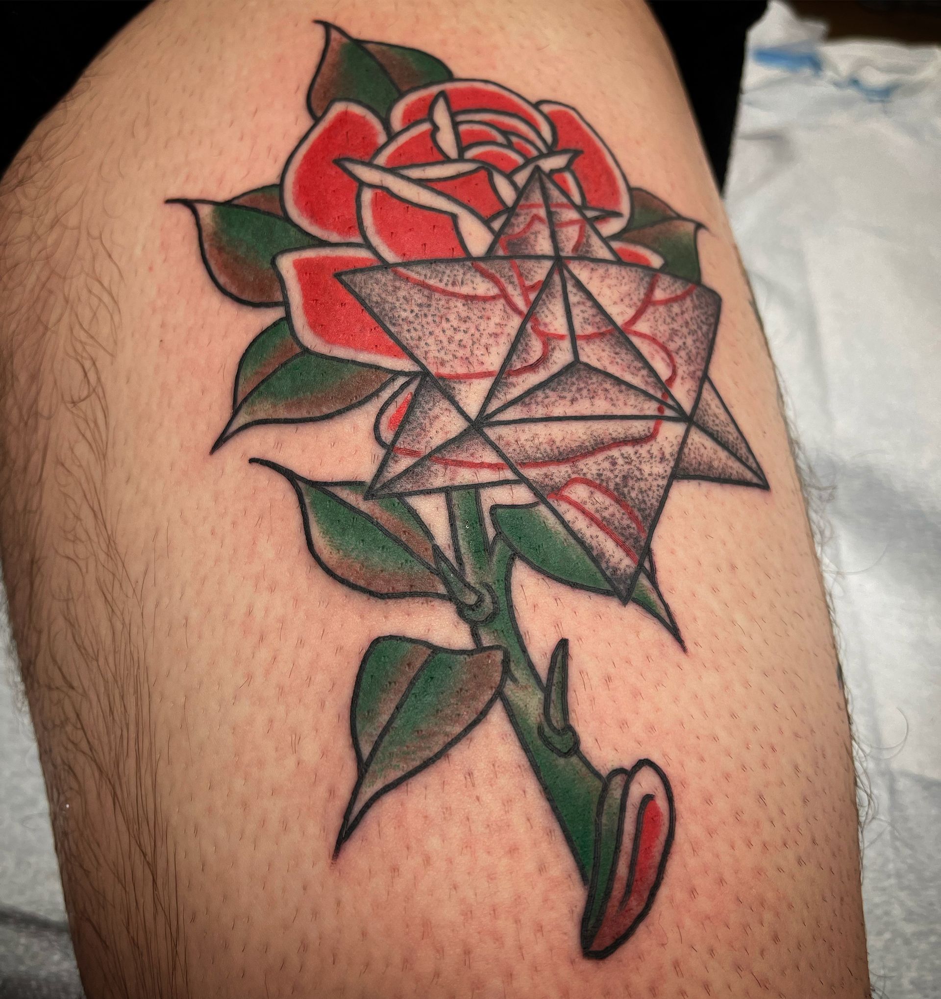 Tattoo of a red rose and a Merkaba star on a leg, green stem with leaves, black and red lines.