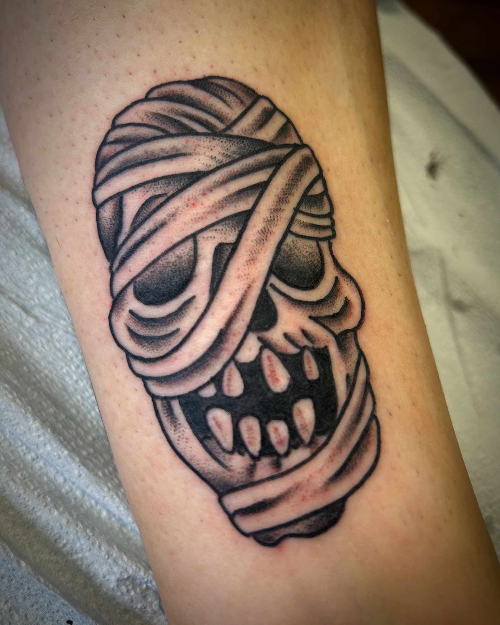 Tattoo of a skull wrapped in bandages, in black and gray shading.