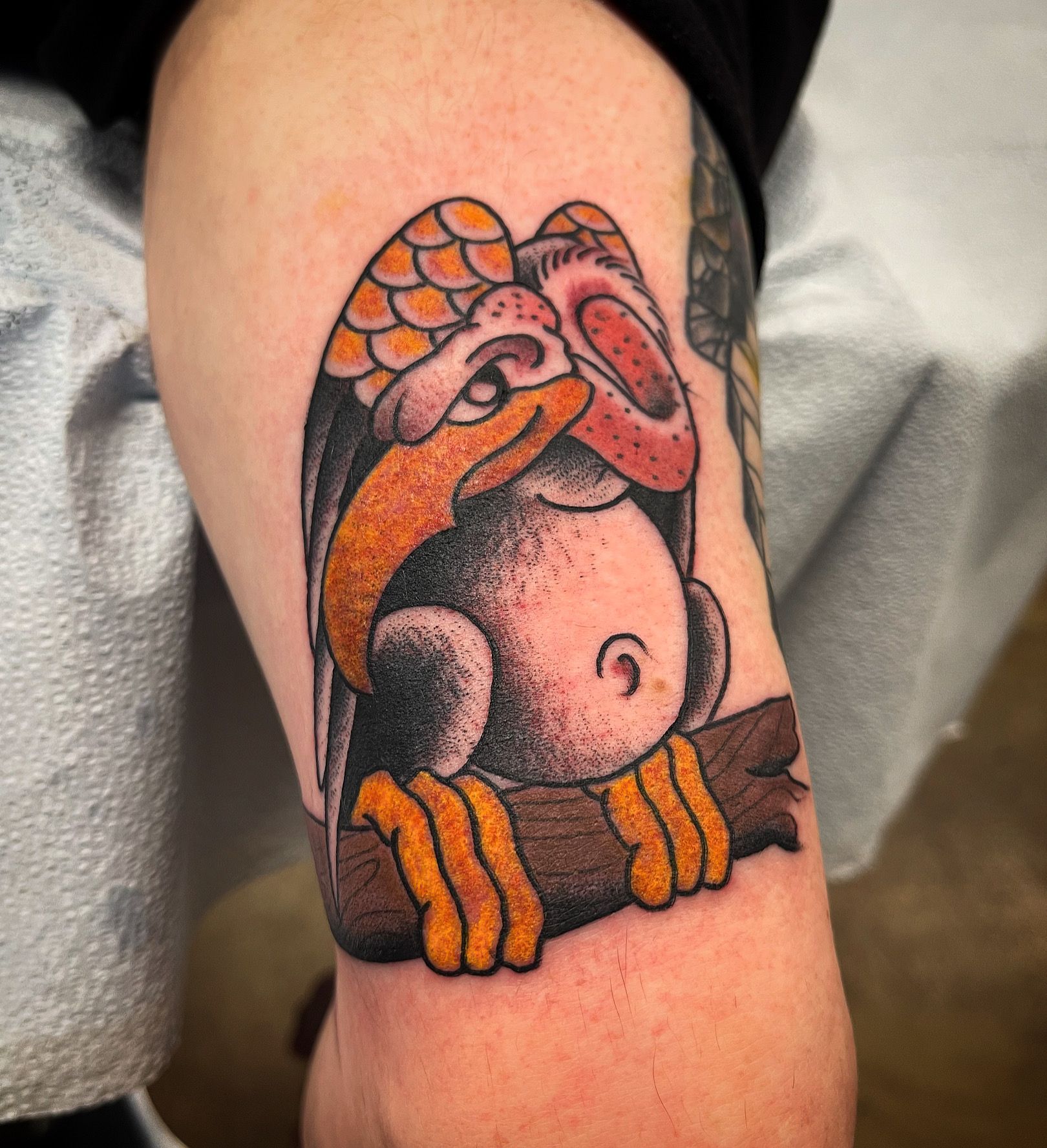 Tattoo of a cartoon vulture with orange beak, feet, and wing accents, perched on a branch.