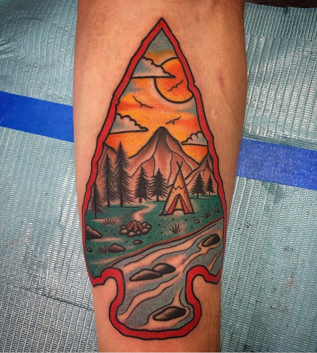Tattoo of a mountain scene within an arrowhead shape, featuring a sunset, trees, a river, and a teepee.