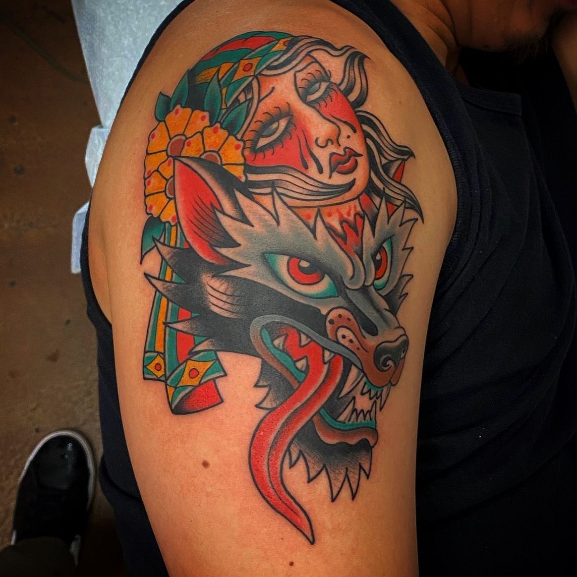 Tattoo of a wolf with red eyes, tongue out, and a woman's face above, on a person's upper arm; colorful.