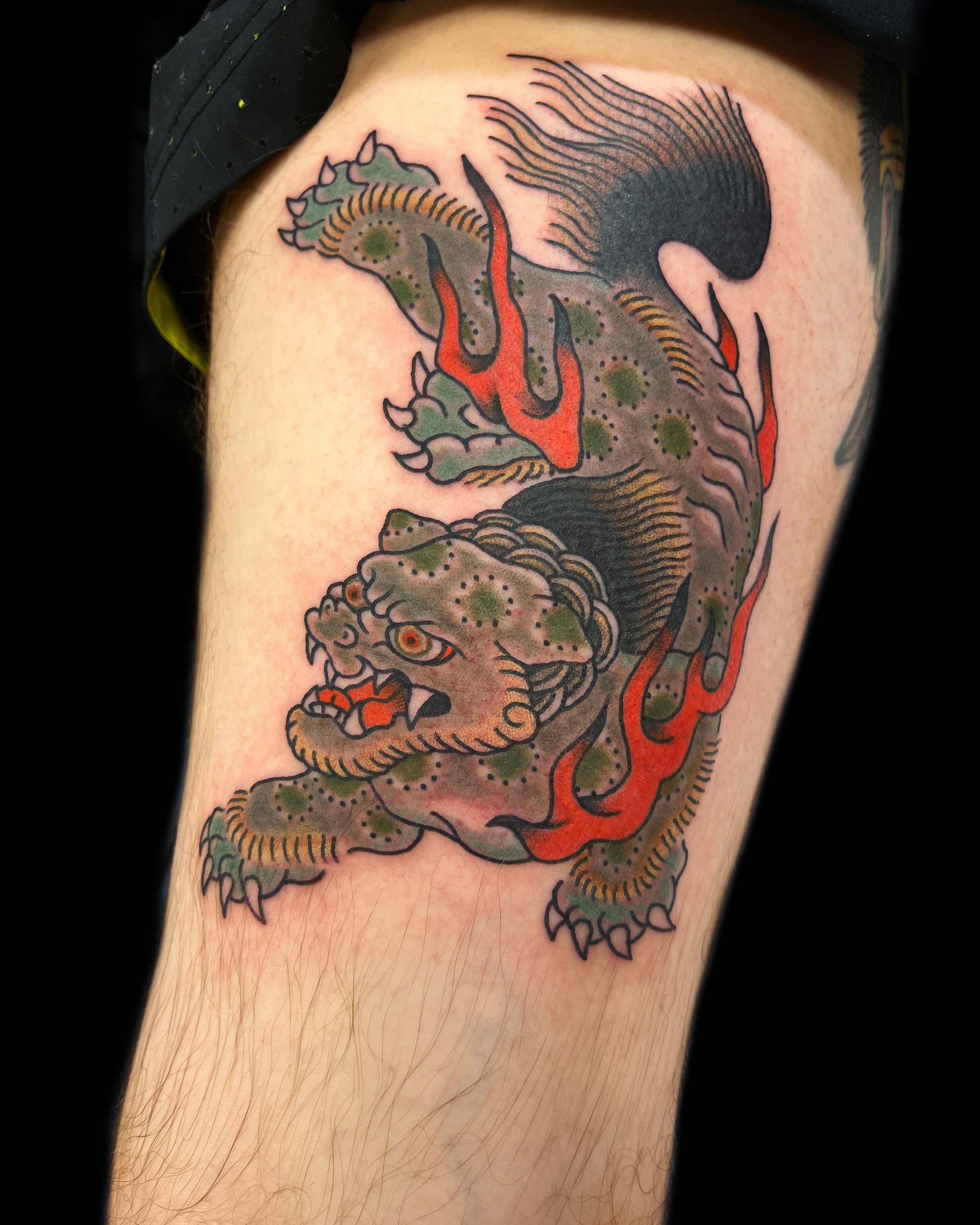 Tattoo of a green mythical creature with orange flames on a person's thigh.