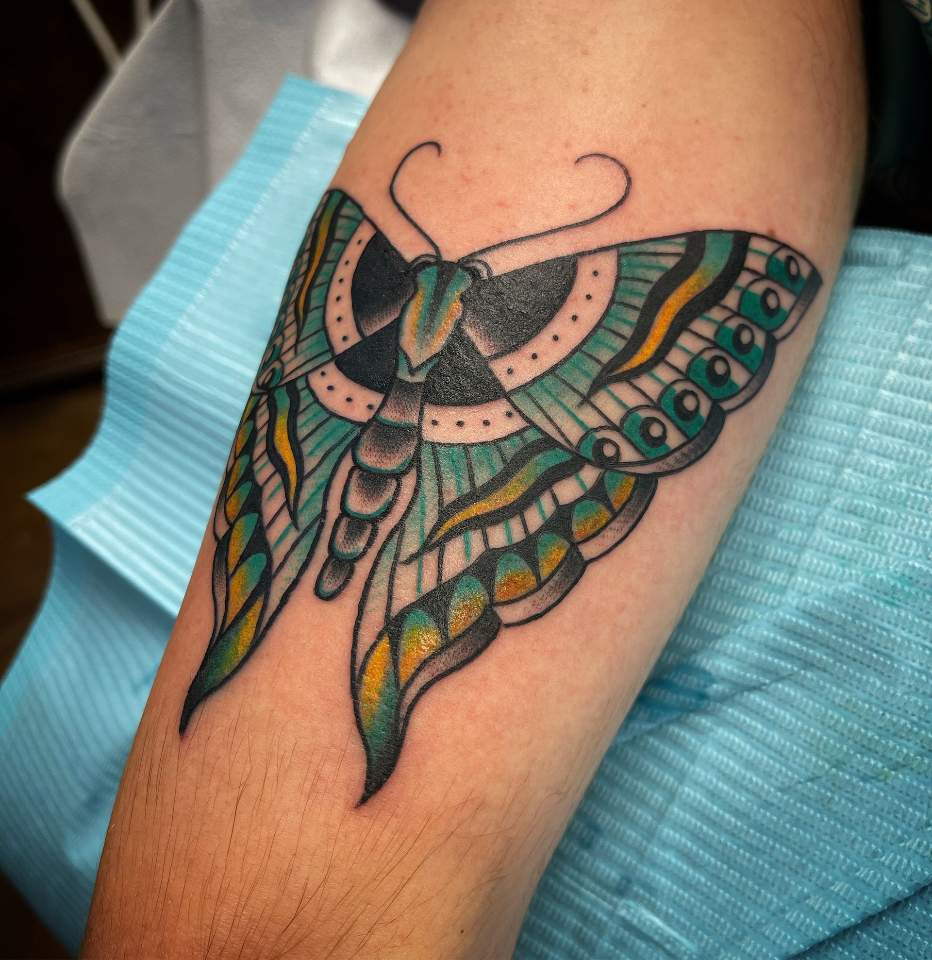 Tattoo of a stylized teal, yellow, and black moth on a person's forearm.