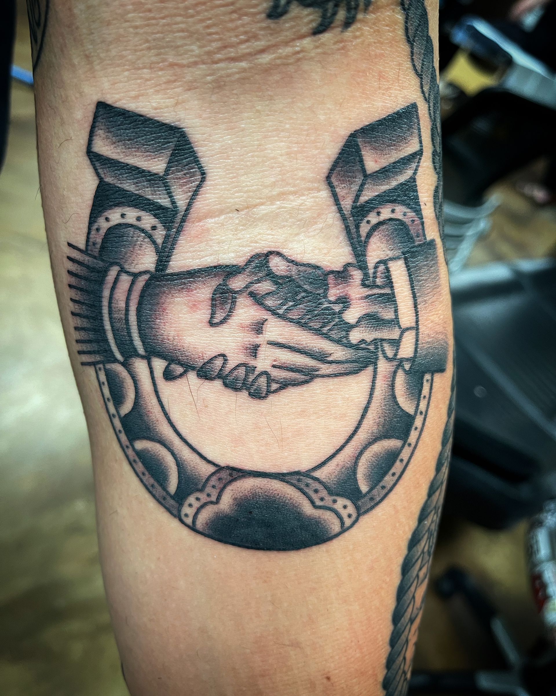 Tattoo of a handshake inside a horseshoe; black and gray with detailed shading on arm.