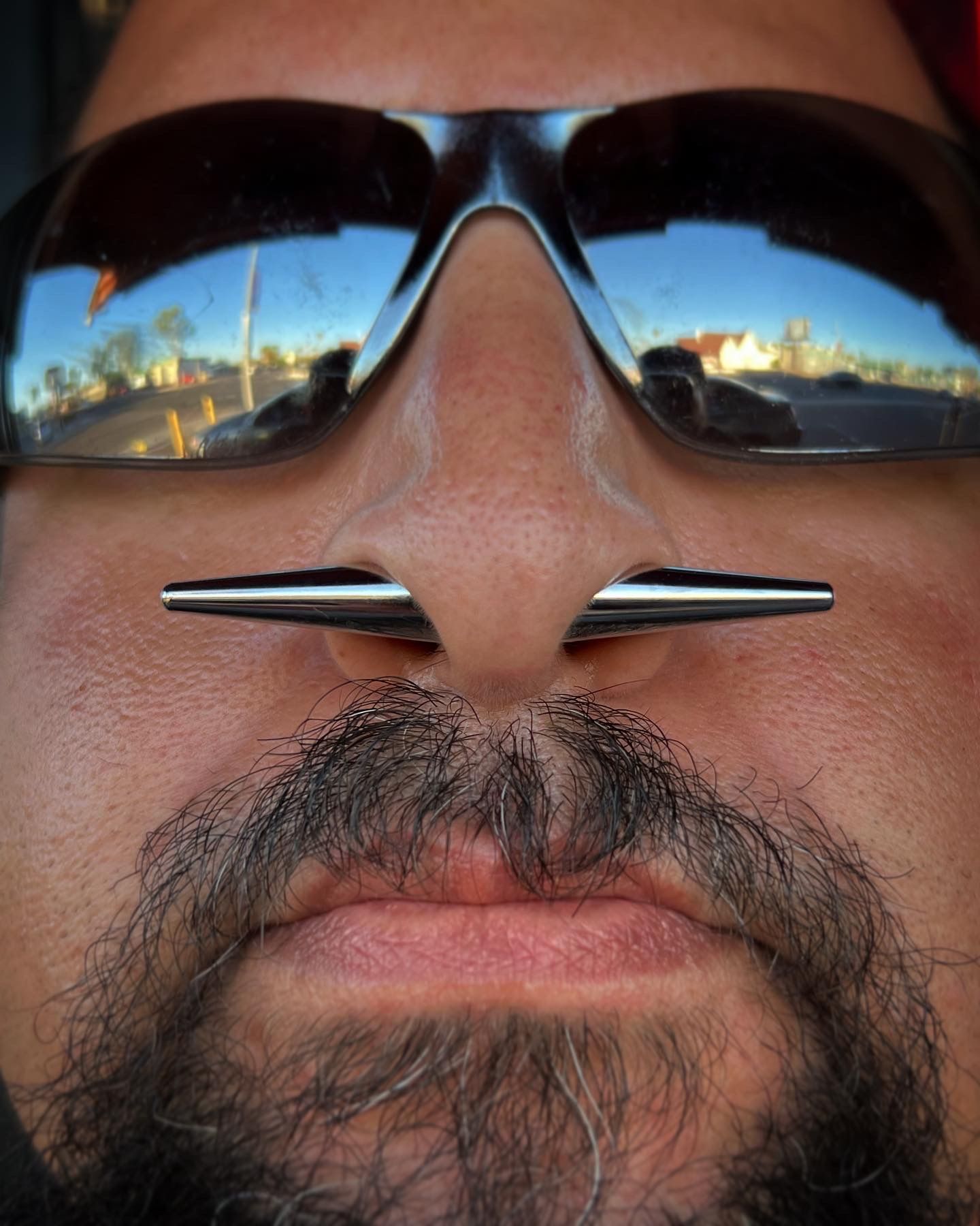 Close-up of a man wearing sunglasses and a silver nose ring, with a city skyline reflected in the lenses.