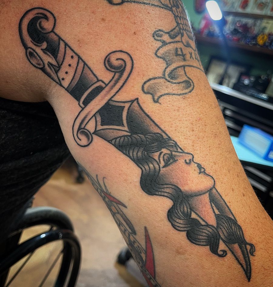 Tattoo of a dagger piercing a woman's face, black and gray ink on an arm.