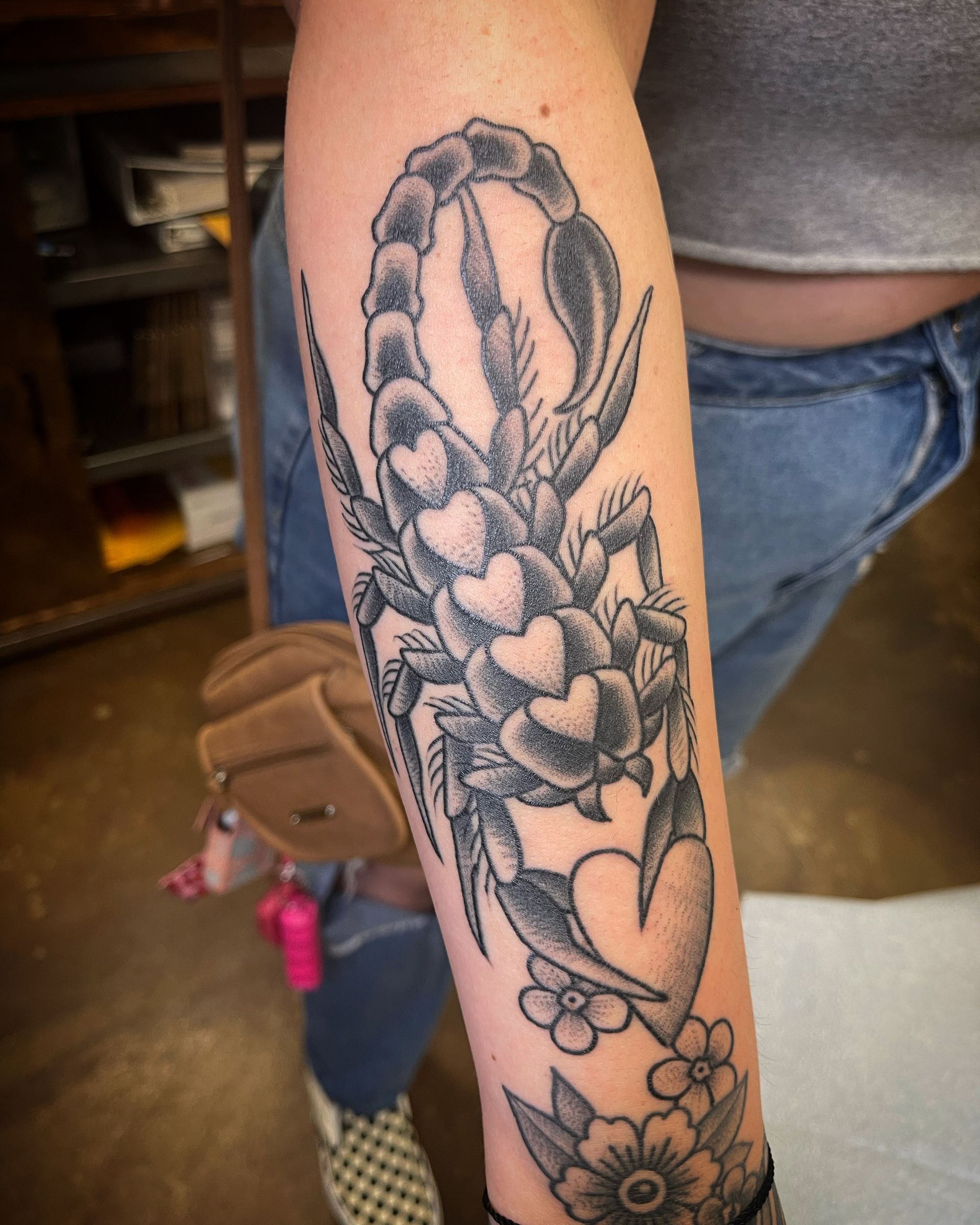 Black and gray scorpion tattoo on an arm, with hearts, flowers, and greenery.