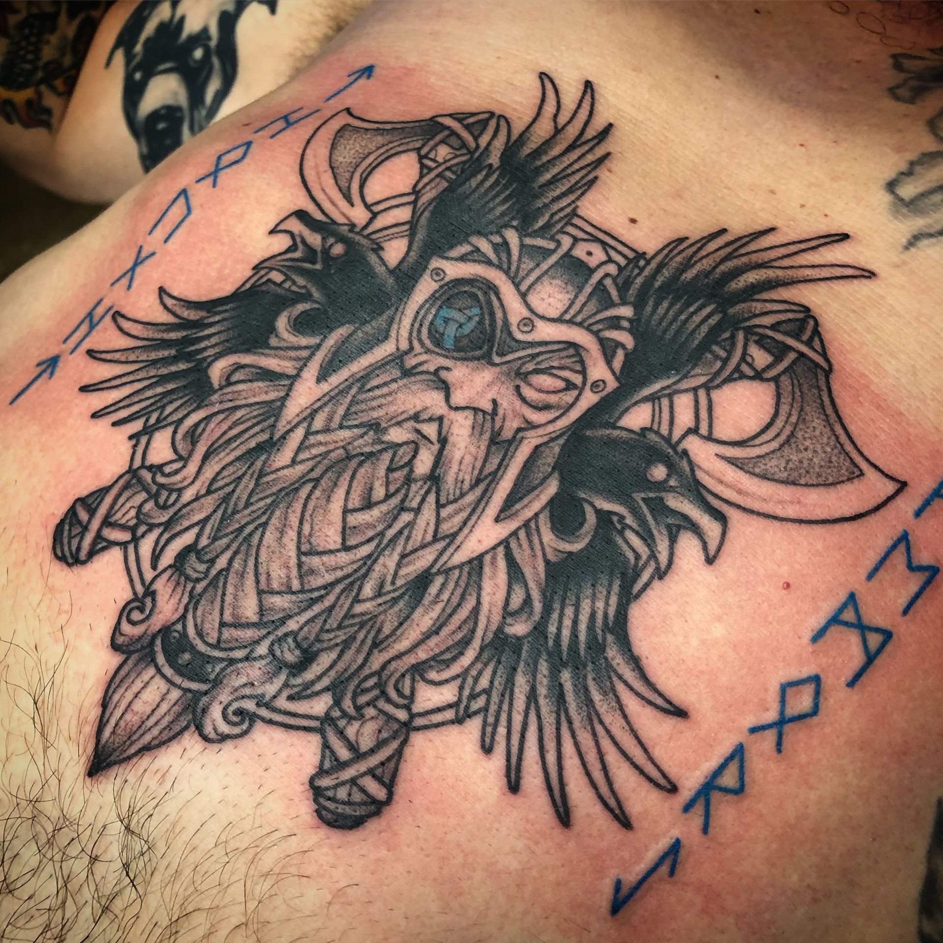 Detailed black ink tattoo of Odin on a chest, with ravens, axes, and rune symbols in blue.