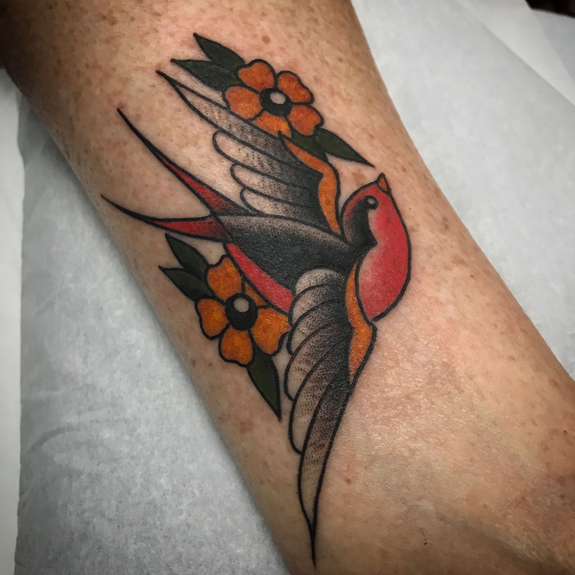 Tattoo of a swallow with red and black body, surrounded by orange flowers and green leaves on an arm.