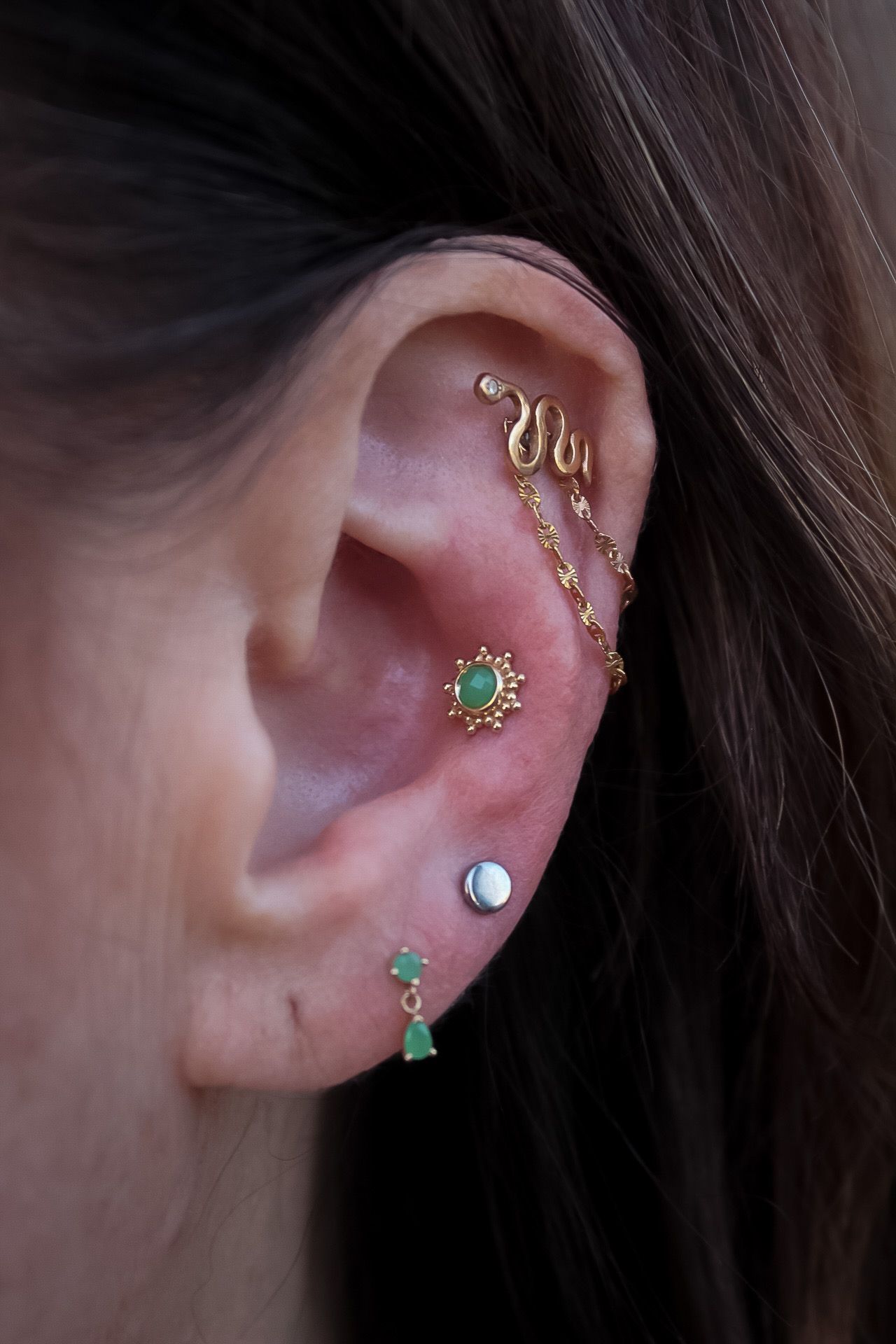 Close-up of an ear with multiple gold and green earrings and chain jewelry