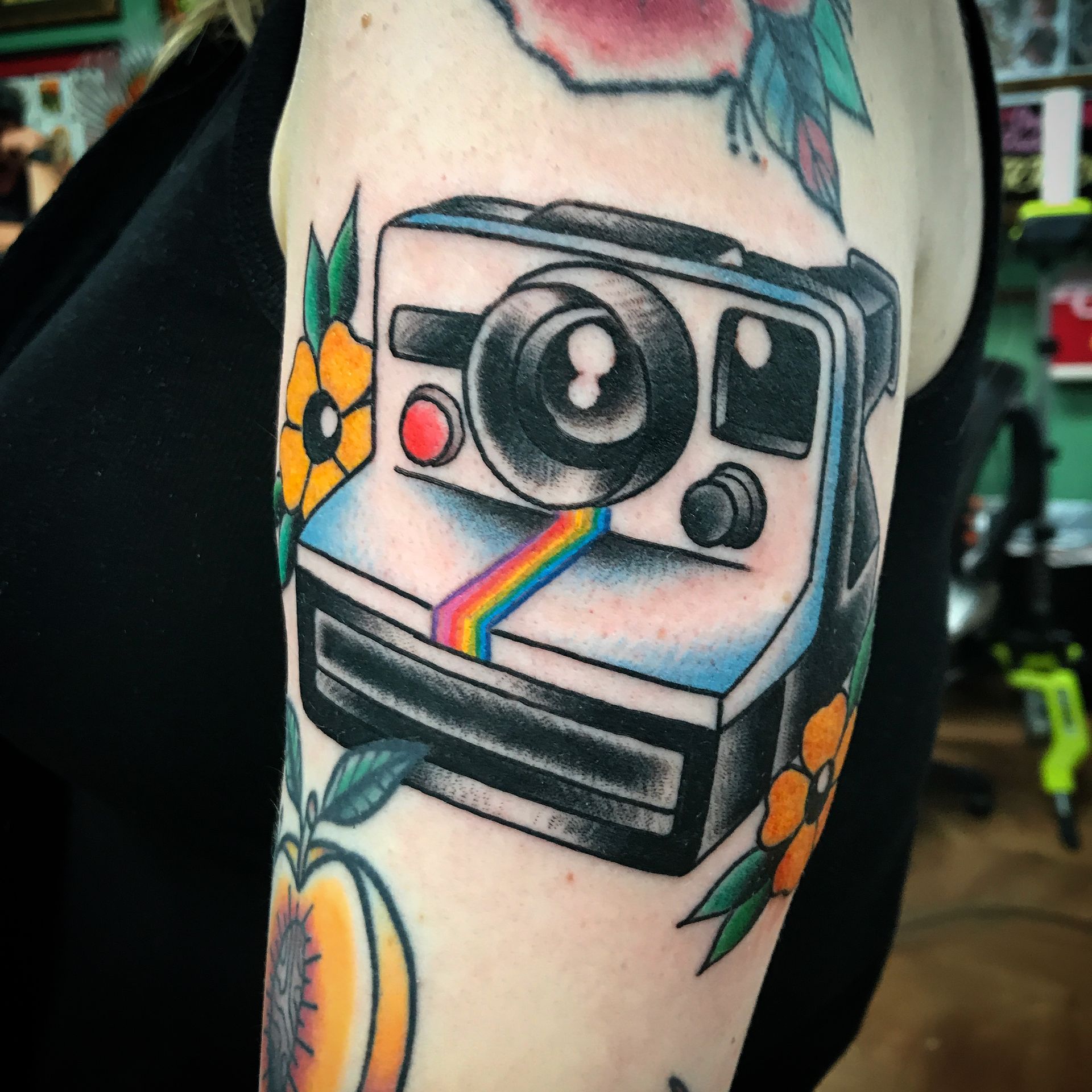 Tattoo of a retro Polaroid camera with a rainbow stripe on an upper arm, surrounded by flowers.