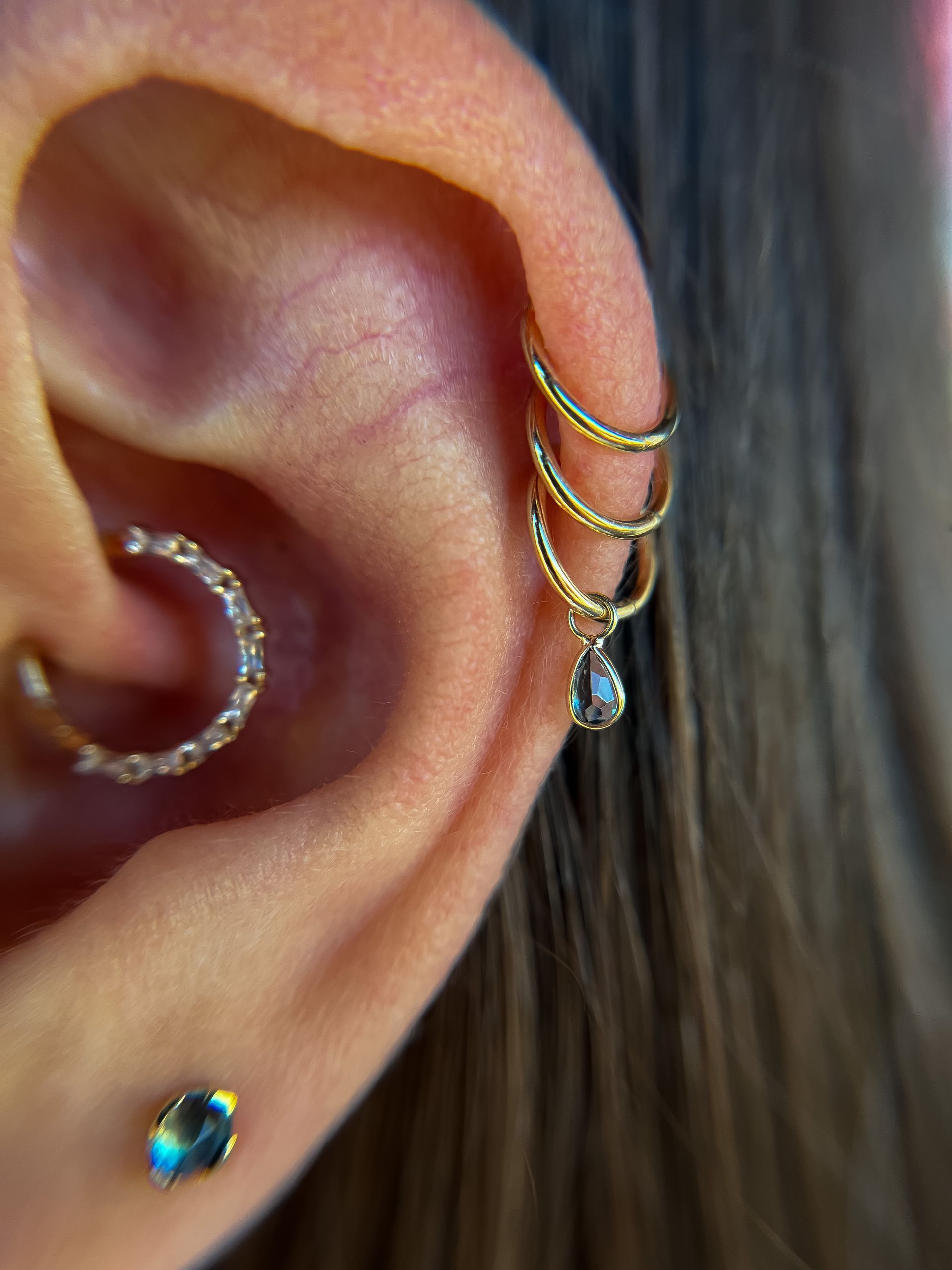 Close-up of an ear with gold hoop and teardrop earrings, plus a small blue stud and ear cuff.