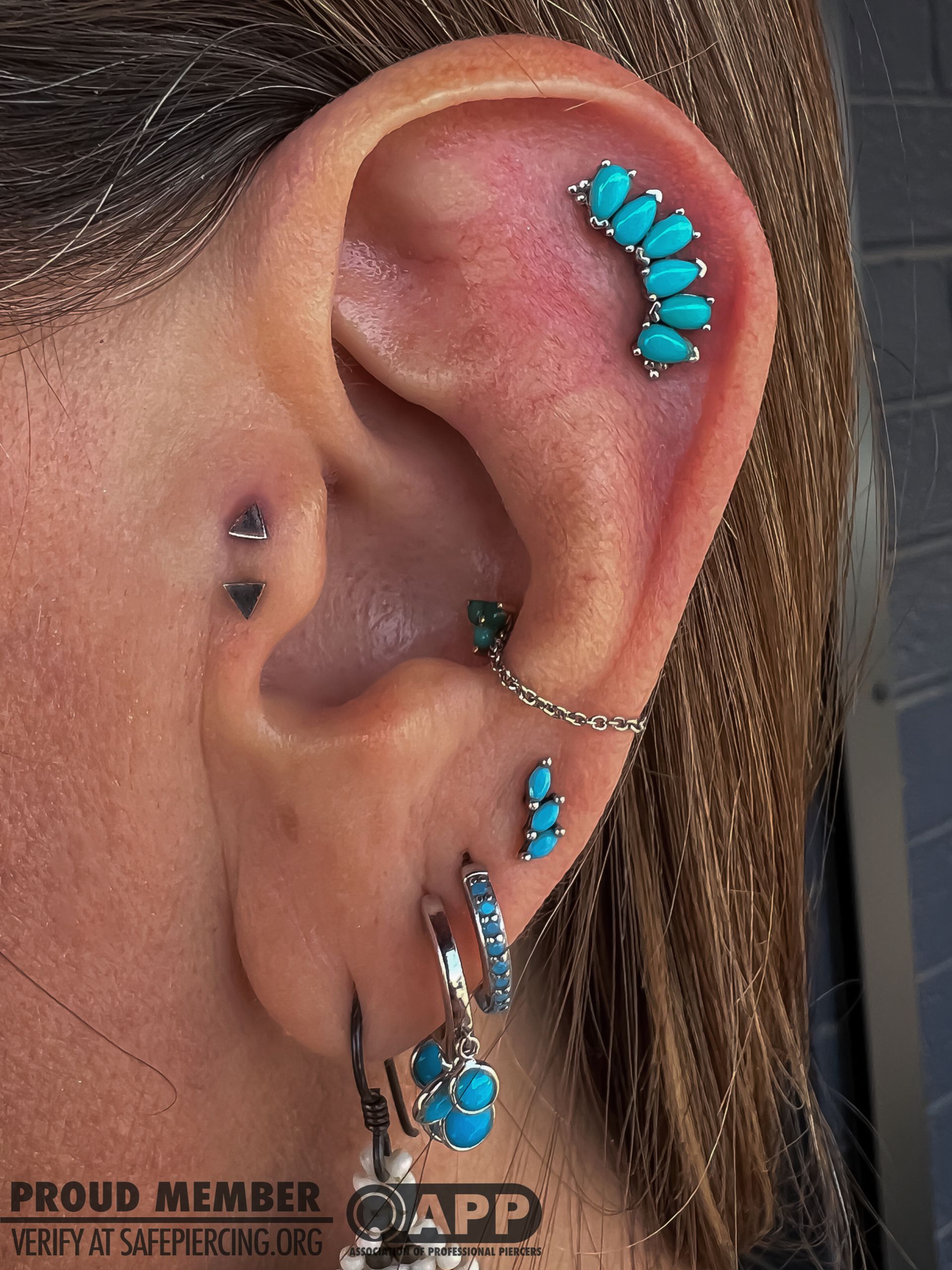 Close-up of an ear with multiple turquoise and silver piercings and a chain earring