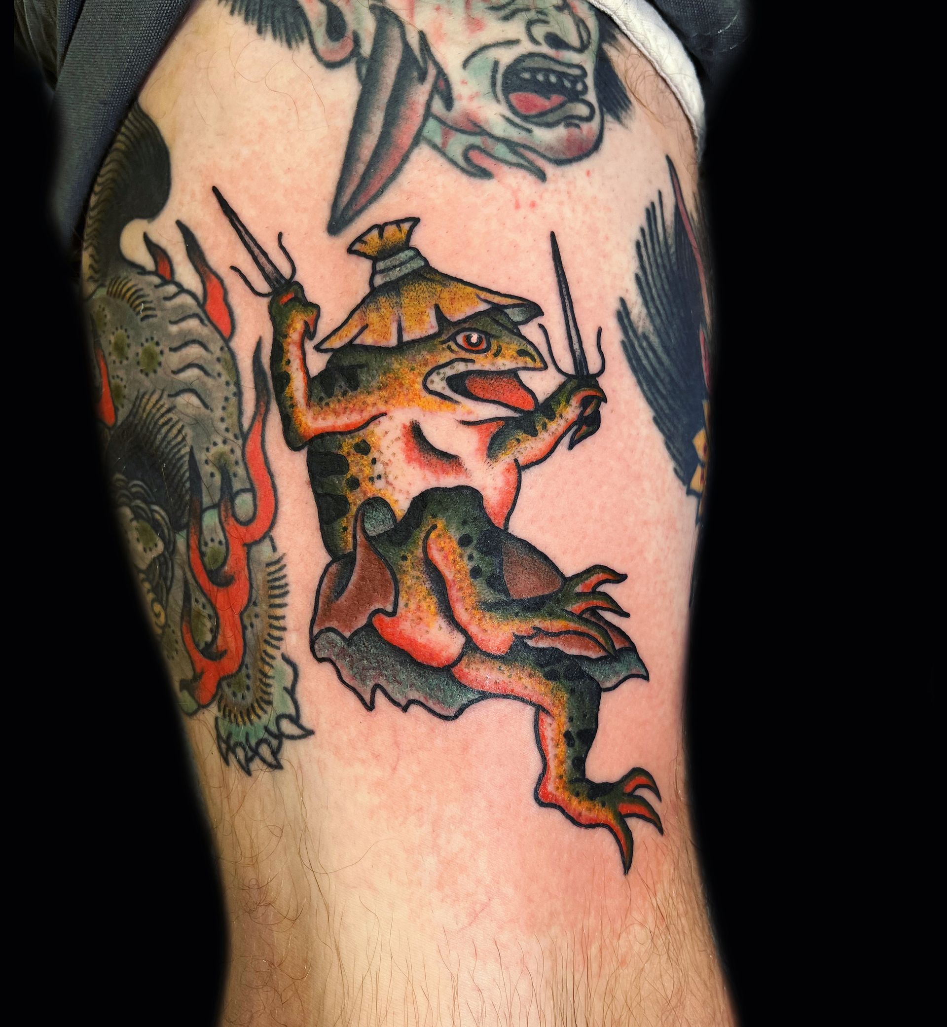Tattoo of a frog wearing a hat, holding drumsticks, and surrounded by other traditional tattoos.