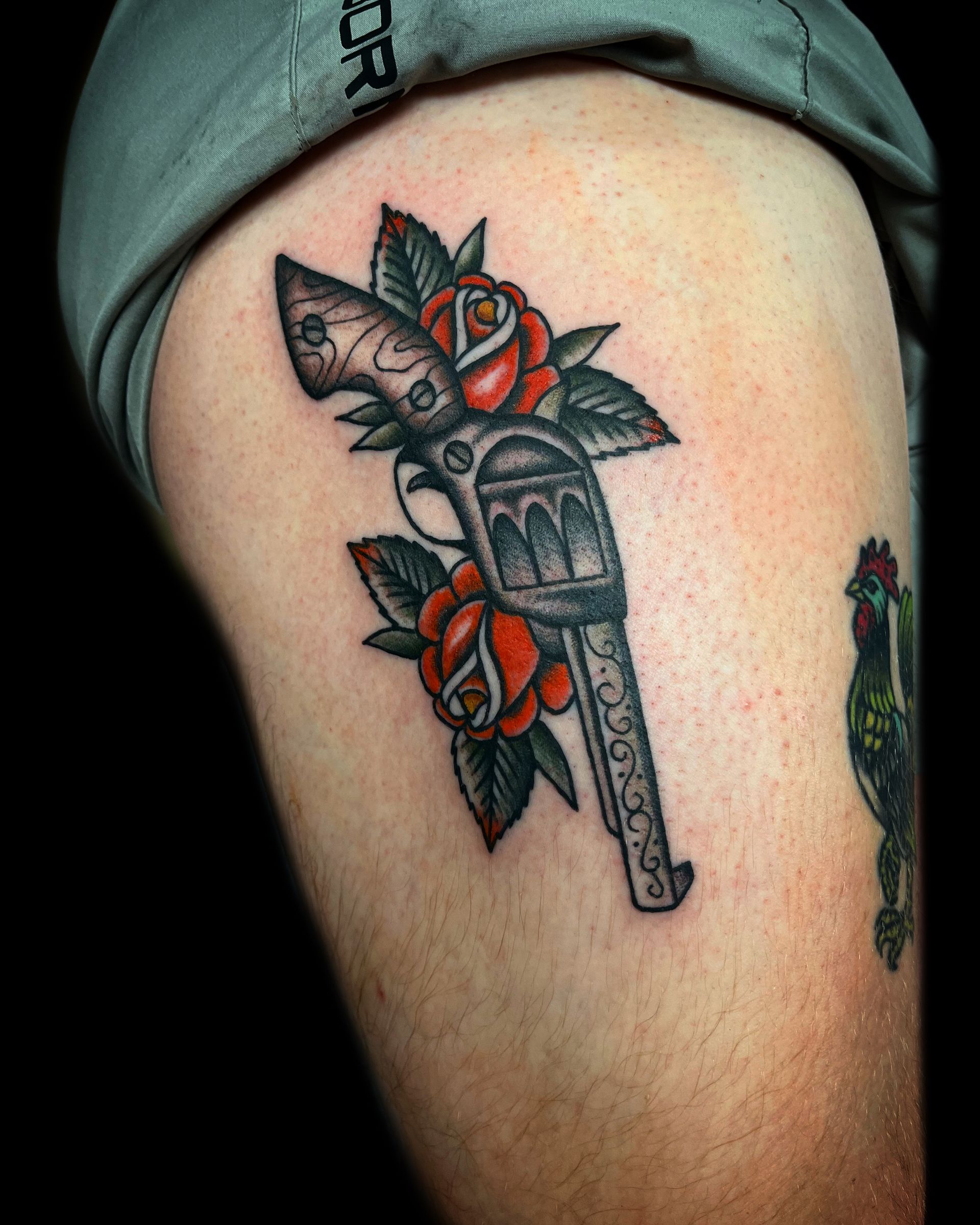 Tattoo of a revolver with red roses and green leaves on an arm.