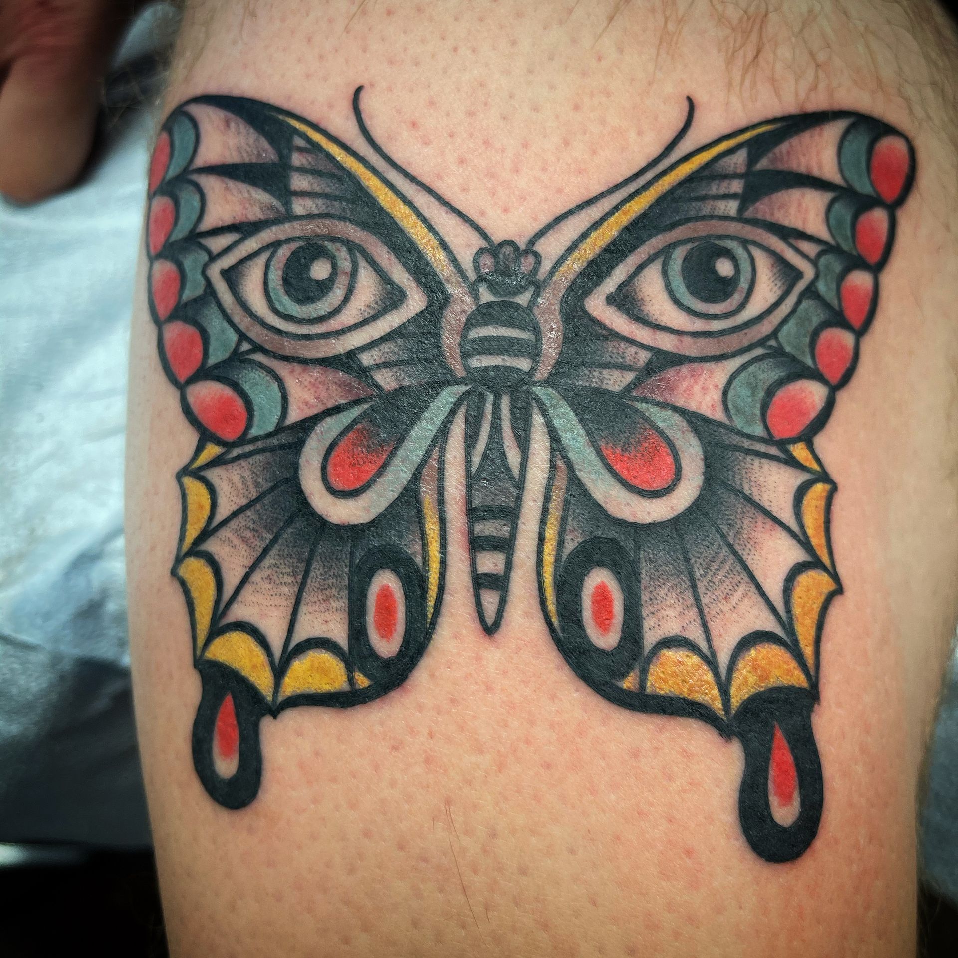 Butterfly tattoo with eyes on a leg. Traditional style, colored with red, yellow, and blue.