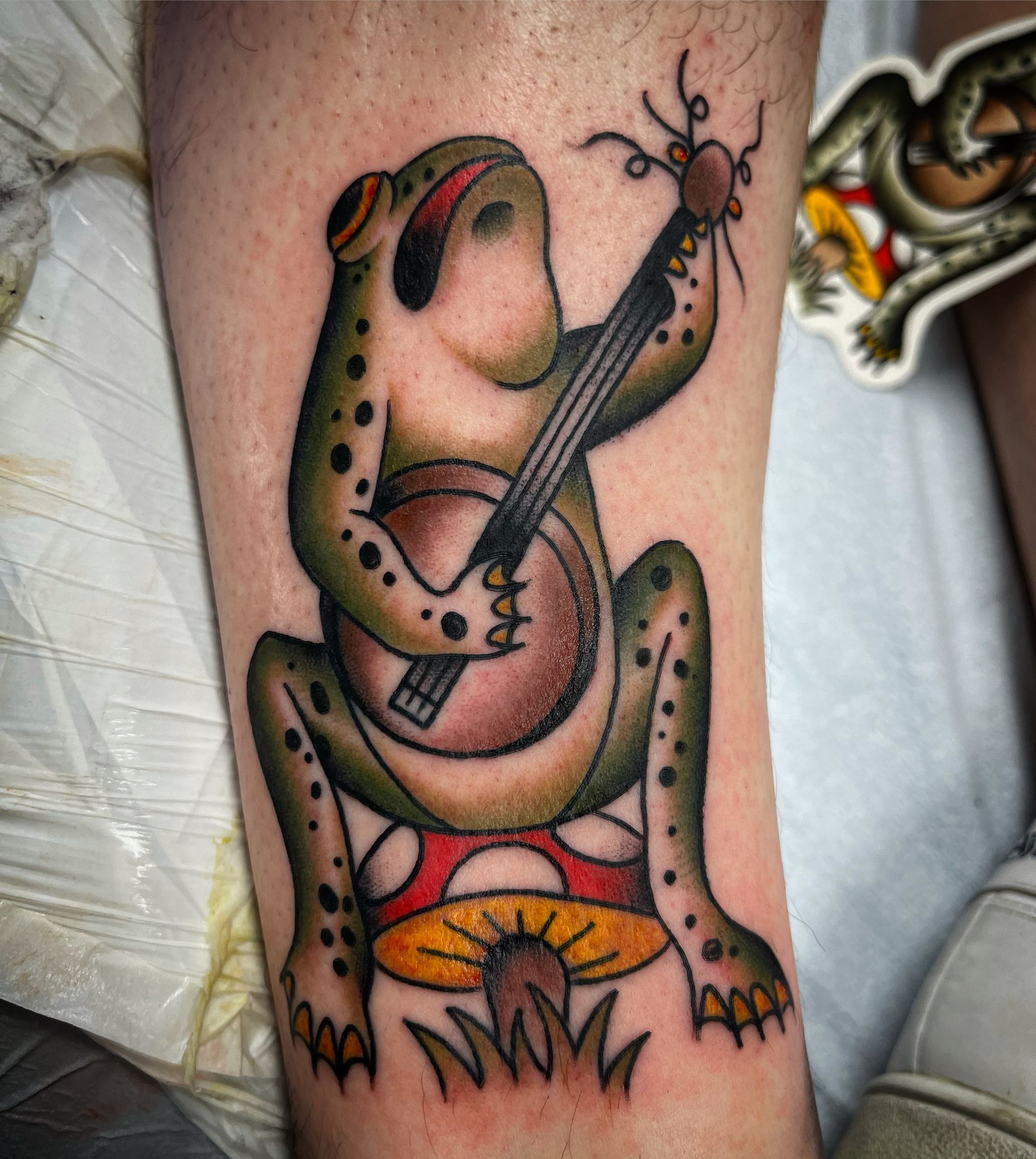 Tattoo of a frog playing a banjo while sitting on a mushroom. Green and brown with black spots.