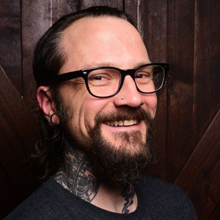A man with a beard and glasses is smiling in front of a wooden wall.