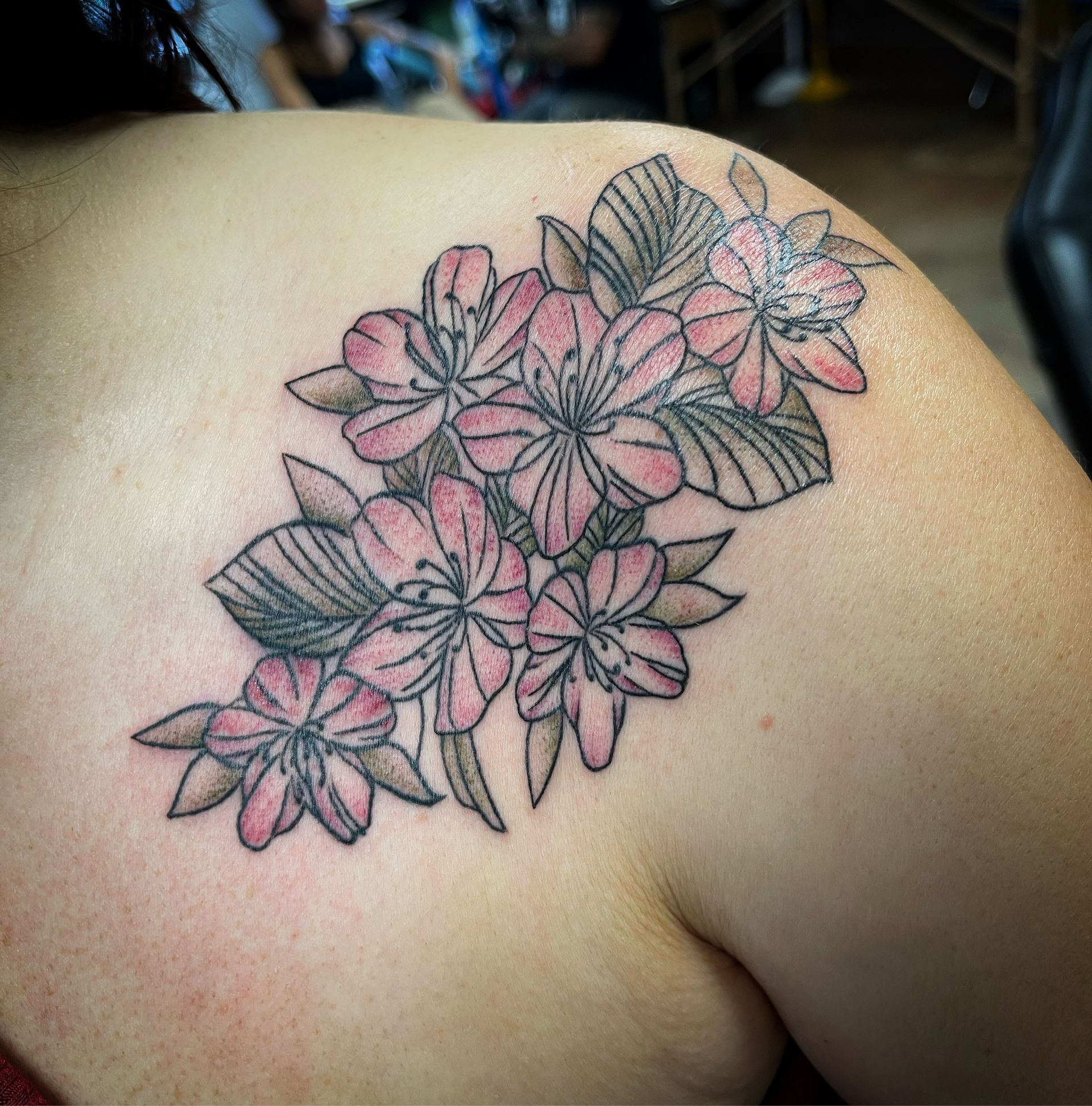 Tattoo of pink flowers and green leaves on a shoulder. Black outline with shaded detail.