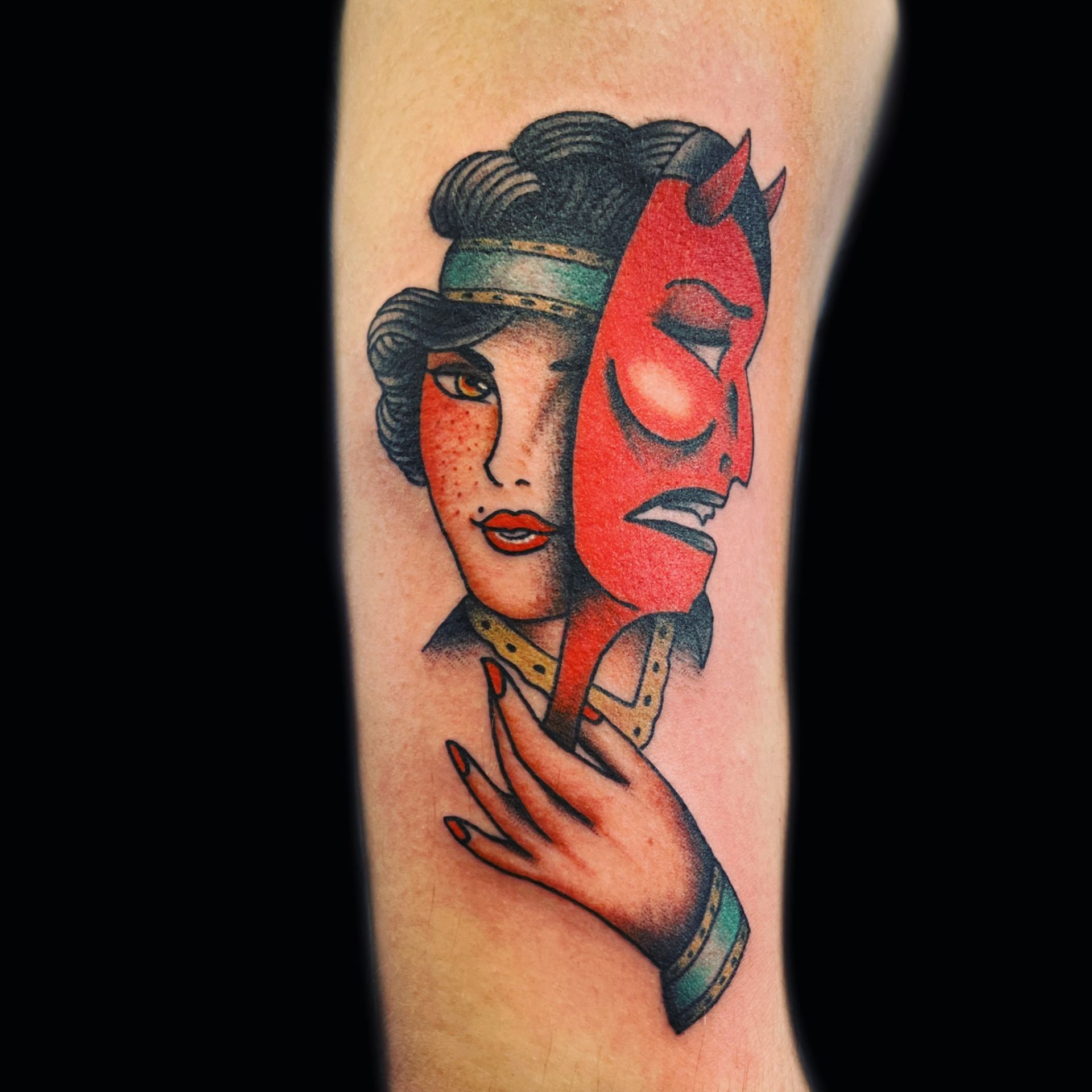 Woman holding a red devil mask, tattooed on an arm. Mask reveals a contrasting emotion to the woman’s expression.