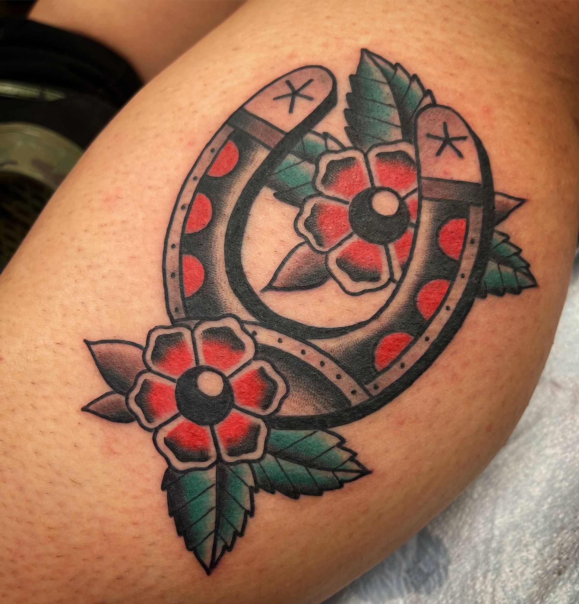 Tattoo of a horseshoe with red and black accents, surrounded by red flowers and green leaves.
