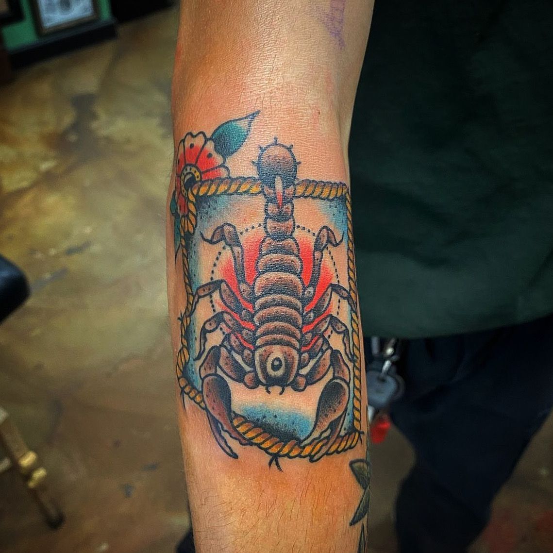 Tattoo of a scorpion inside a rope-framed box on a person's arm, with red and blue coloring.