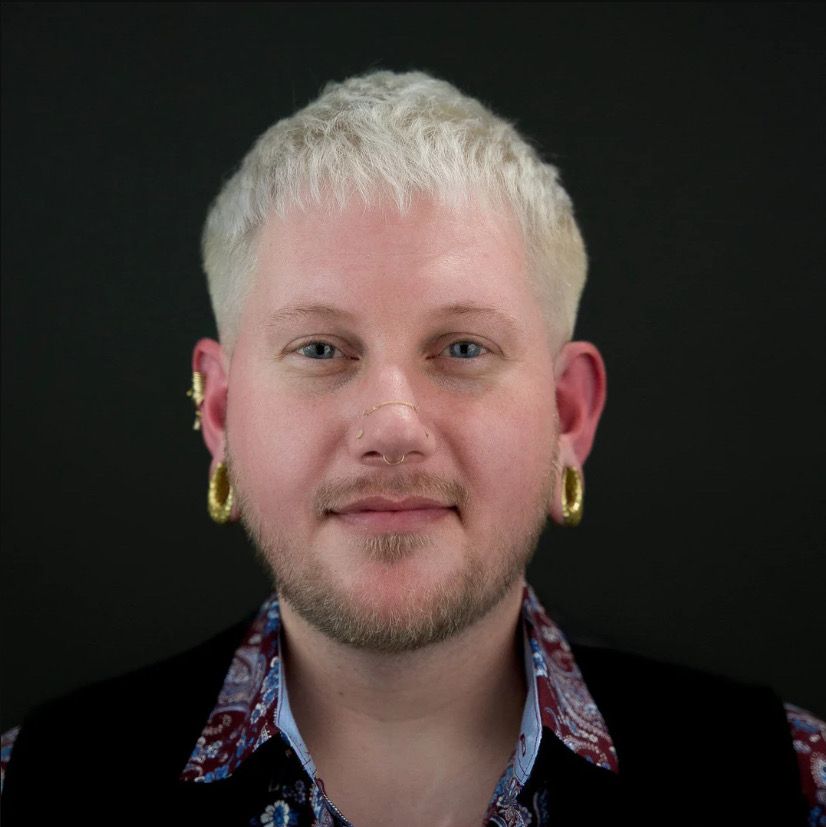 Portrait of a person with platinum blond hair, gold hoop earrings, and a patterned shirt against a dark background