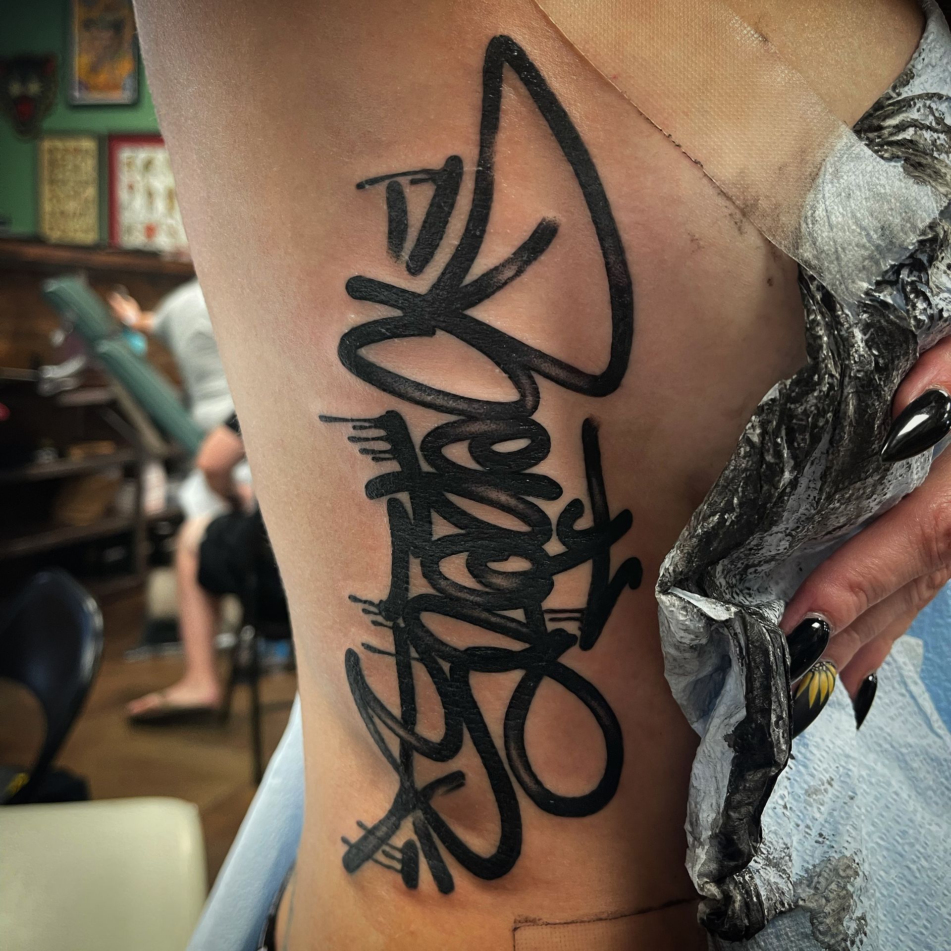 Black graffiti-style tattoo on a person's side; a gloved hand holds a cloth. Tattoo parlor in background.
