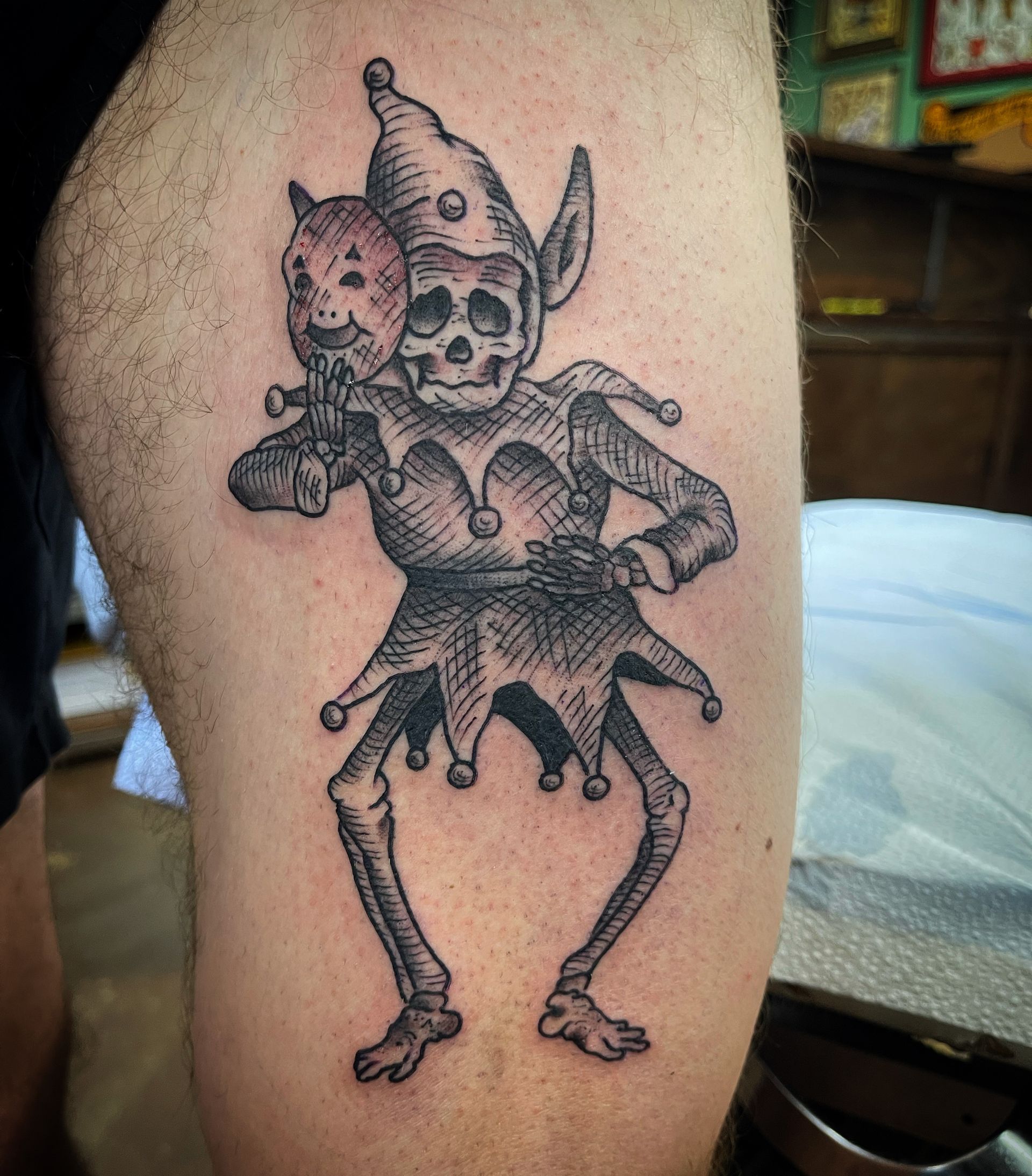 Tattoo of a skeletal jester holding a mask, wearing a hat with bells, on a leg.