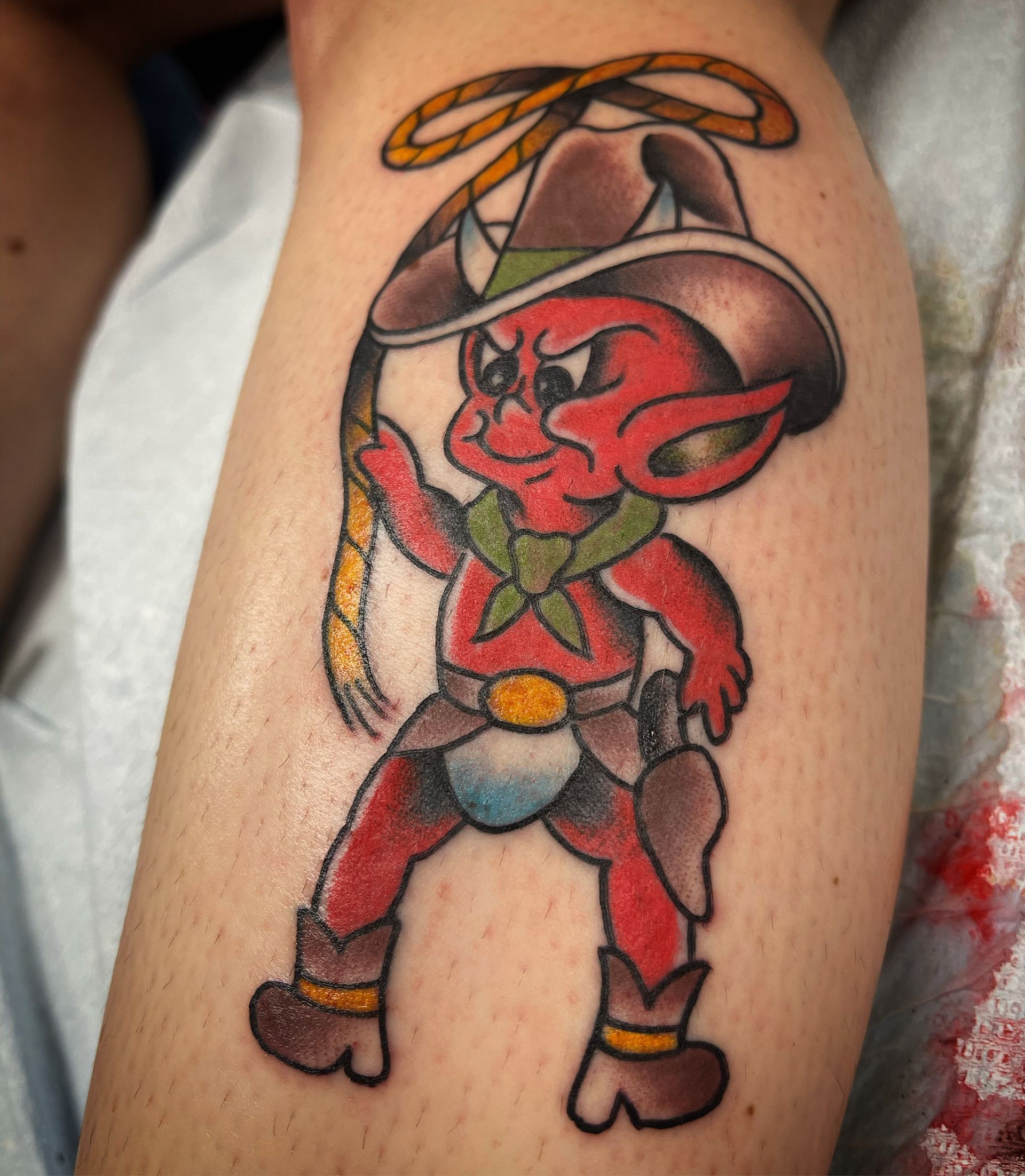Cowboy devil tattoo on leg. Red demon in cowboy hat holds lasso, wearing chaps, boots, and holster.