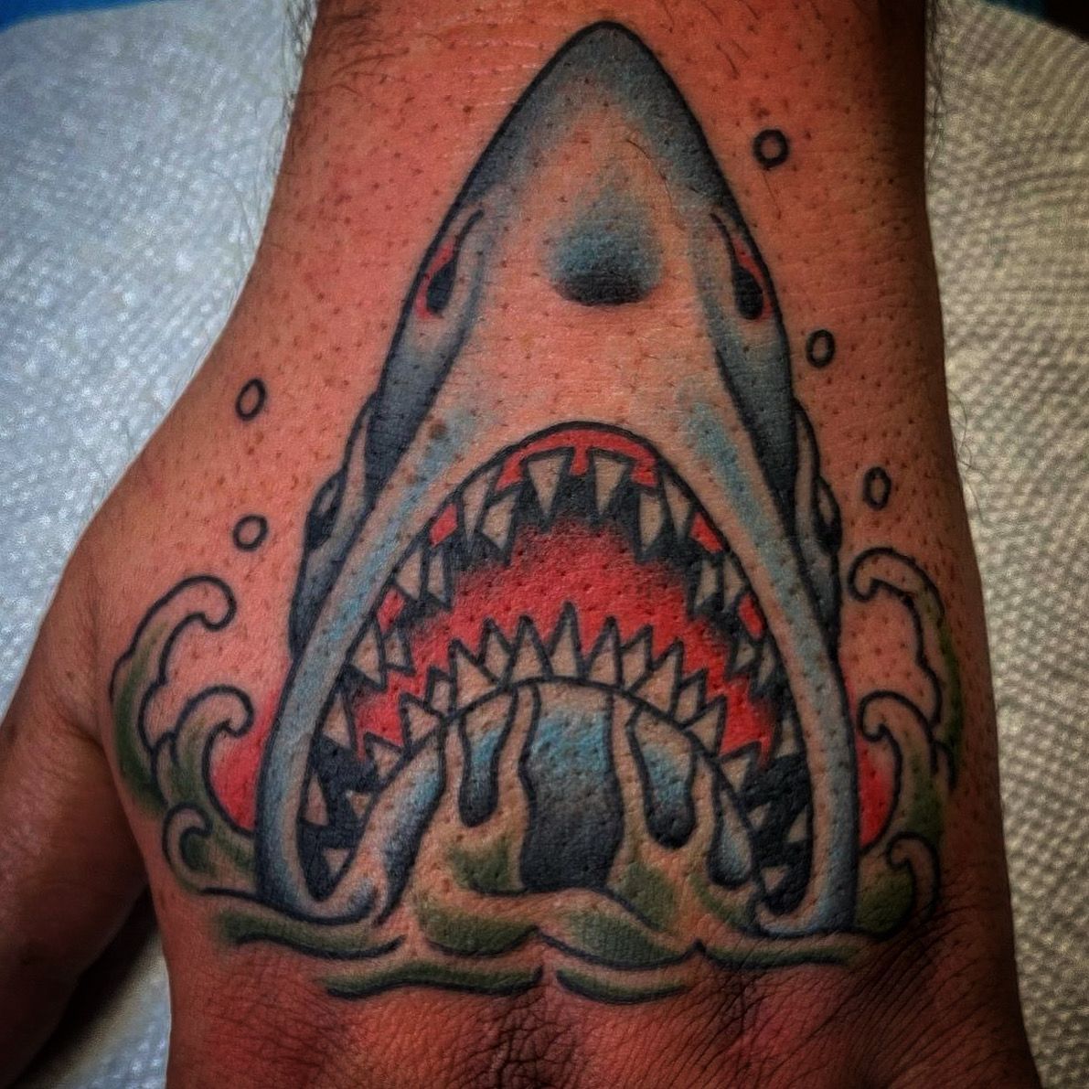 Tattoo of a shark's open mouth with jagged teeth, surrounded by waves, on a hand; blue, red, and green colors.