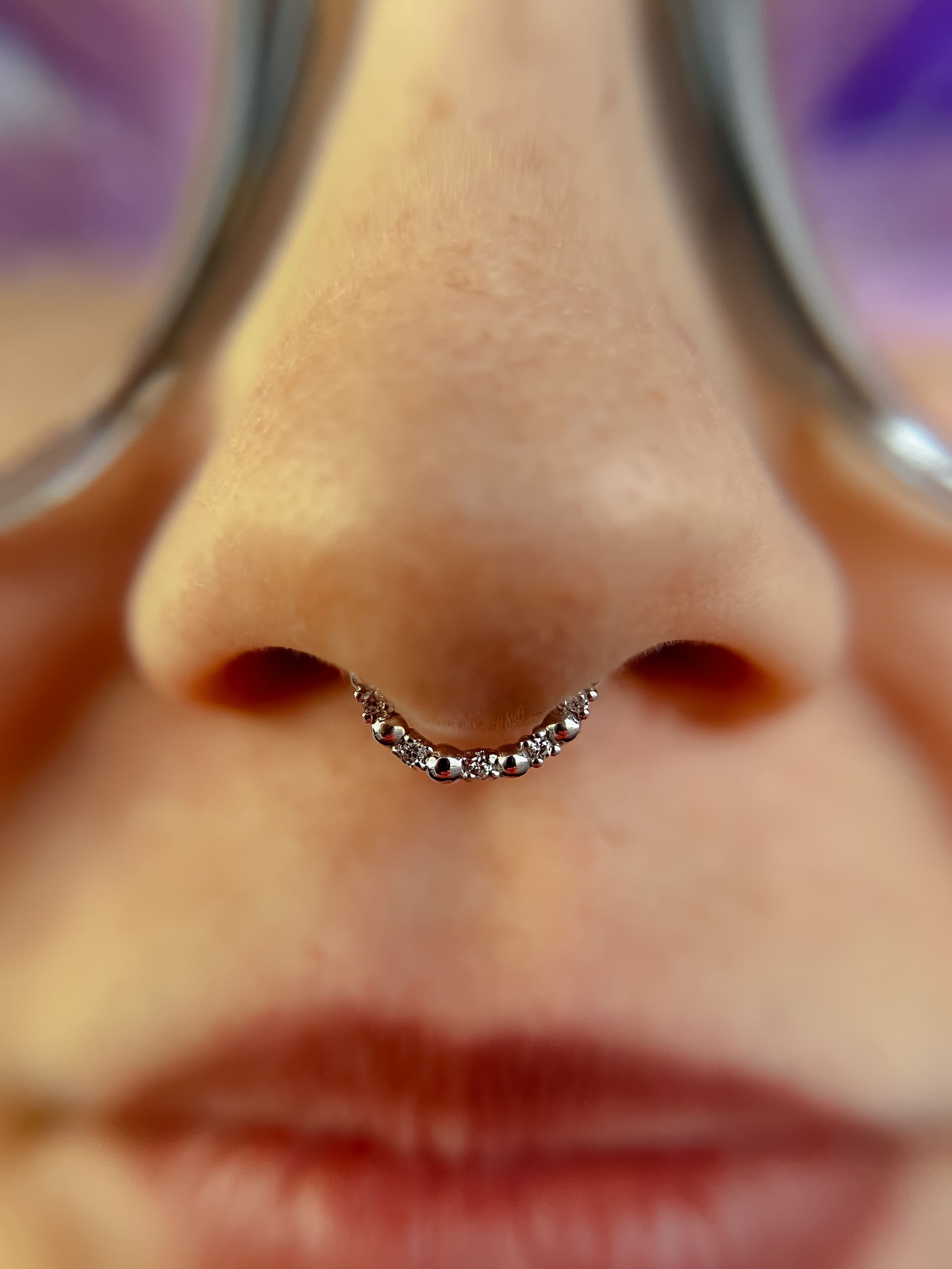 Close-up of a nose with a small silver septum ring and blurred glasses above the lips
