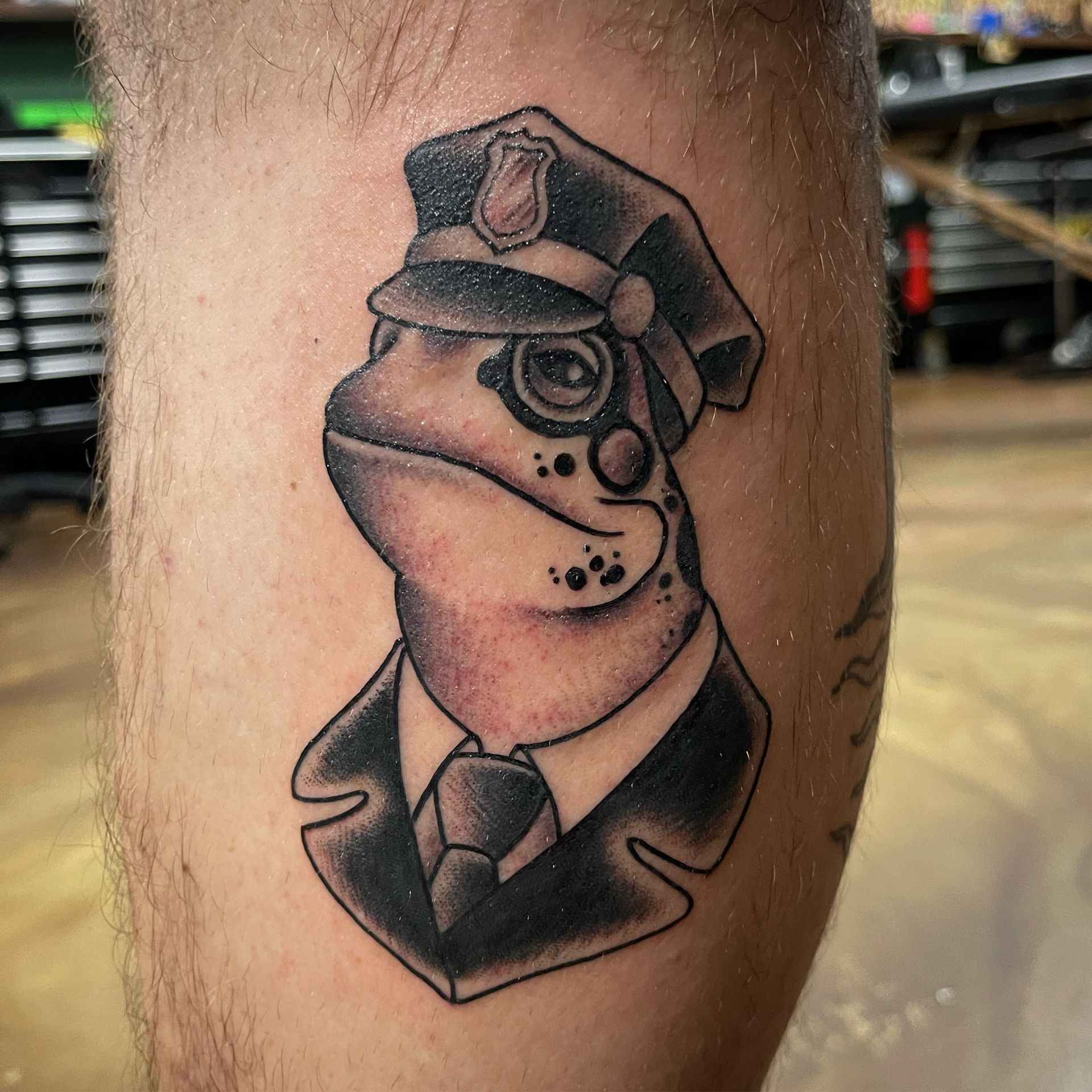 Tattoo of a frog wearing a police hat, uniform and monocle.