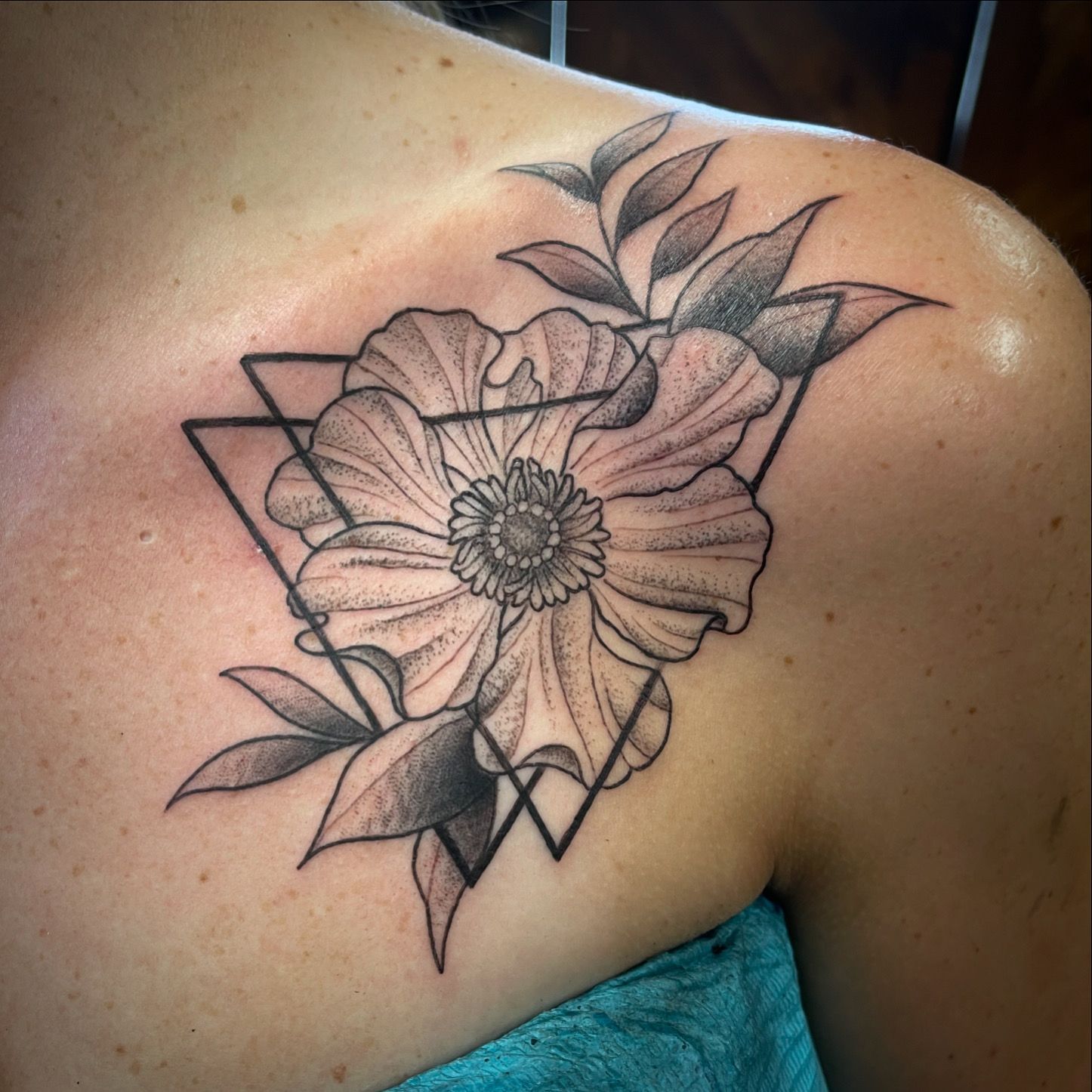Black and white floral tattoo on a shoulder, featuring a flower within a geometric triangle and leaves.