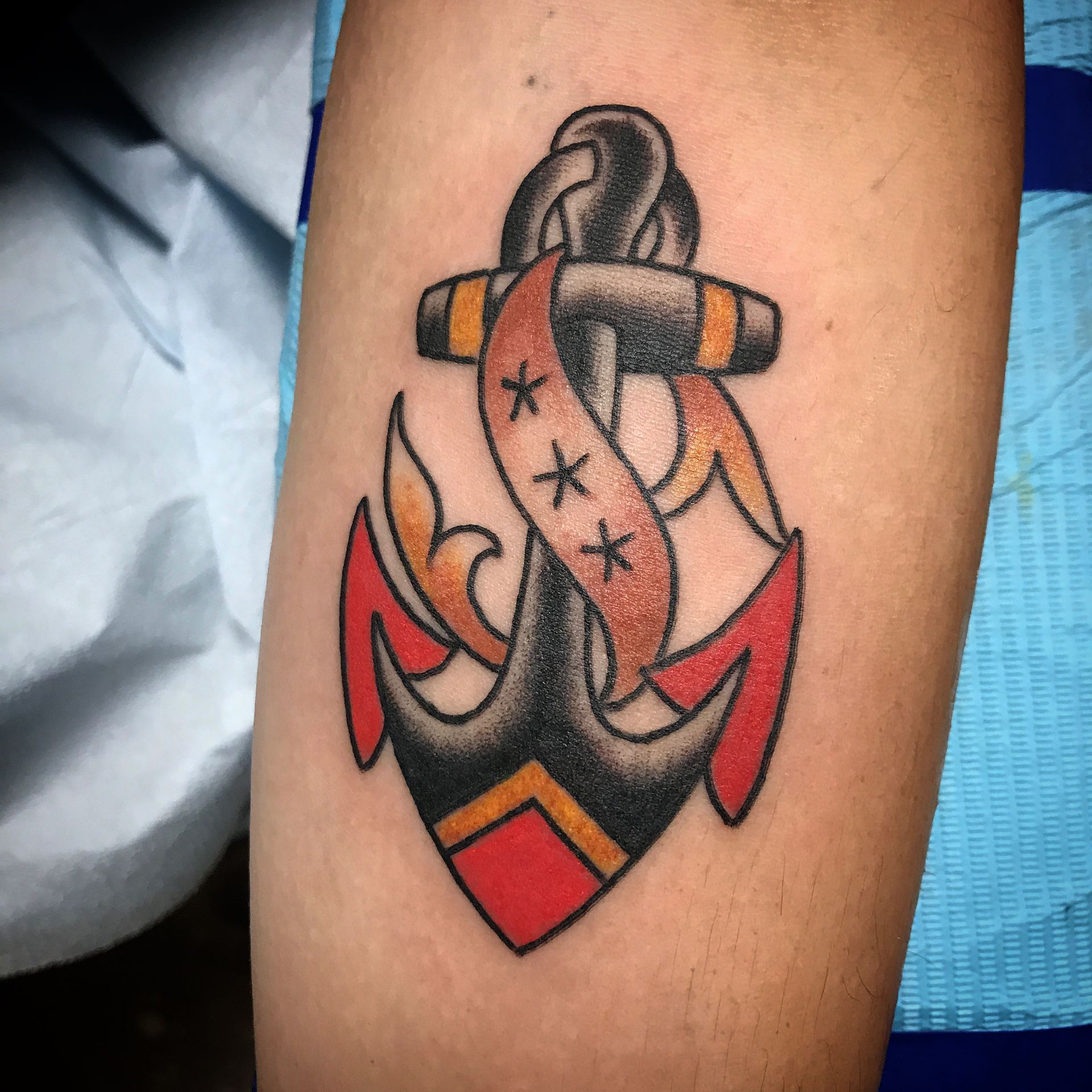 Black and red anchor tattoo with orange ribbon and stars on arm.