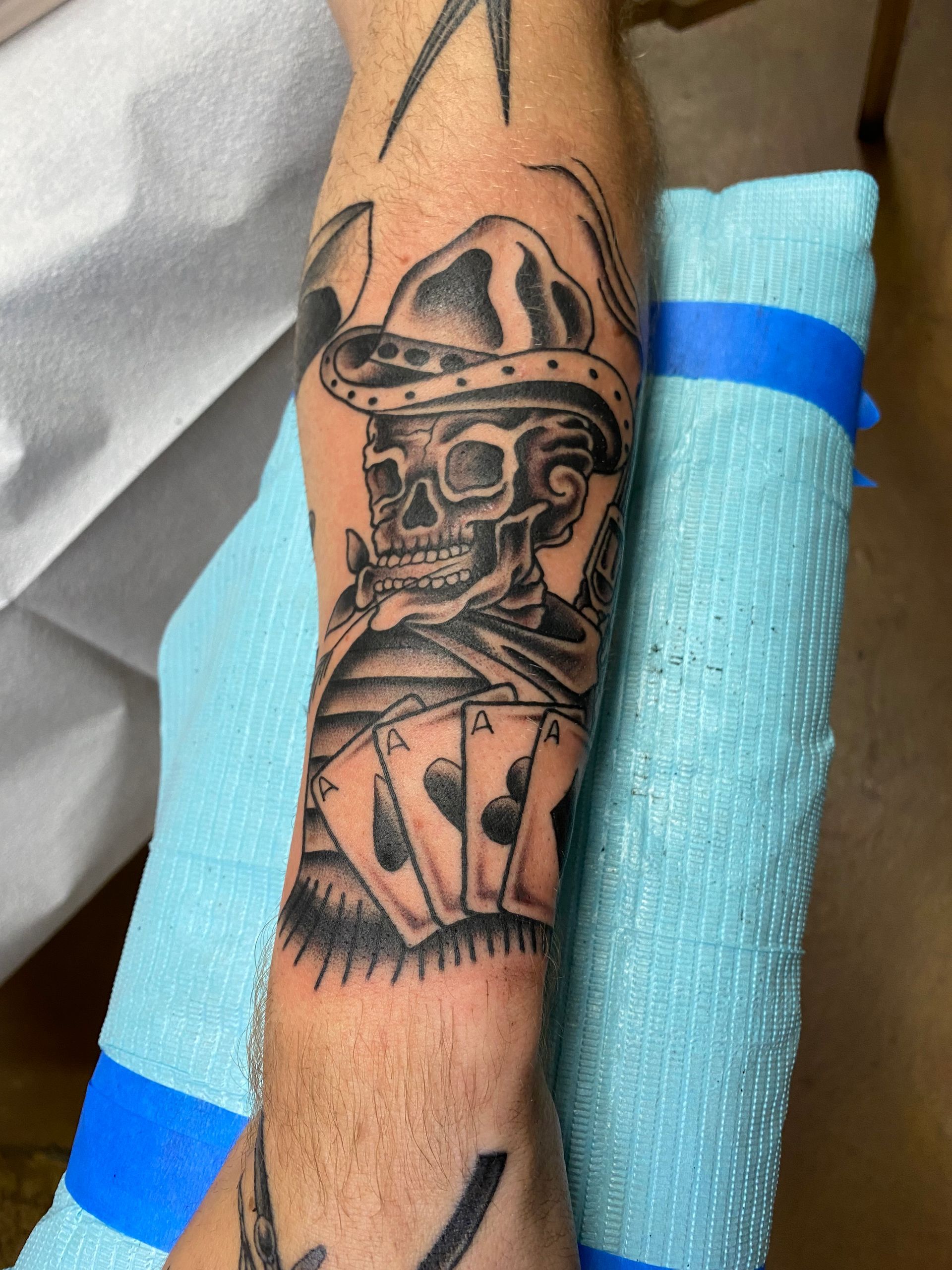 Tattoo of a skull in a cowboy hat holding four playing cards, with a blue bandage.