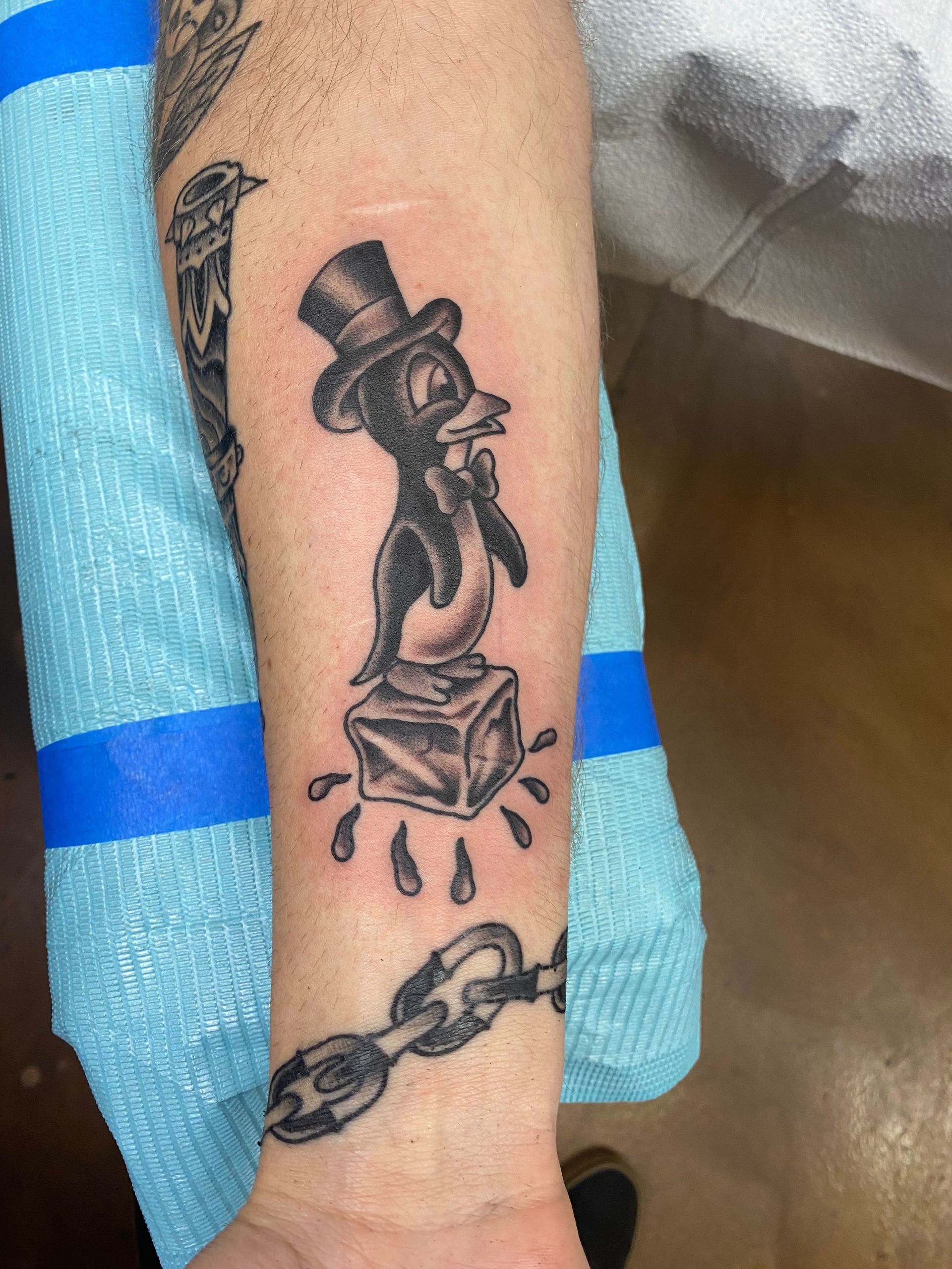 Tattoo of a penguin wearing a top hat atop an ice cube, with a chain link below.