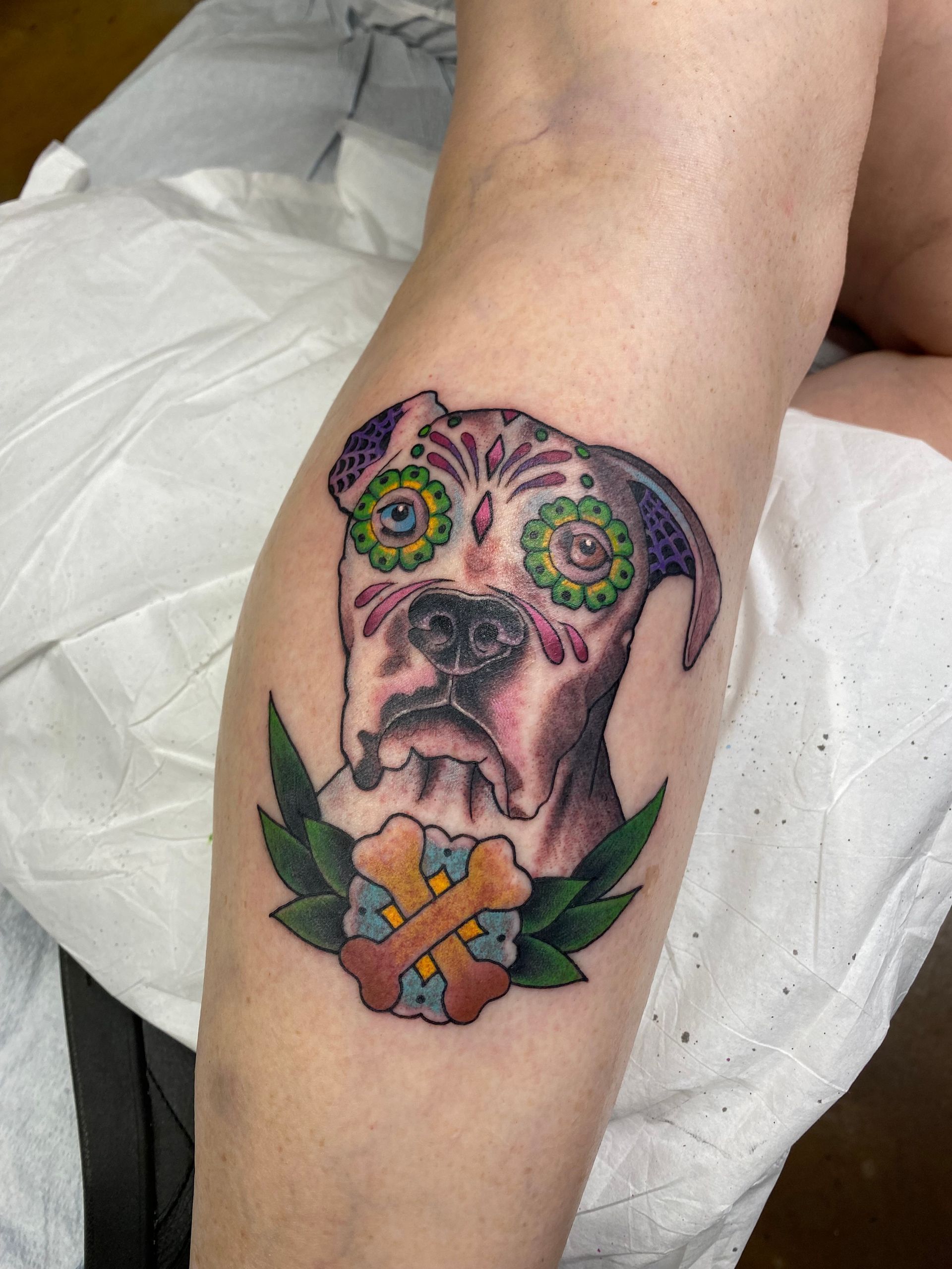 Boxer dog tattoo in Day of the Dead style on a leg, with bone flower and green leaves.