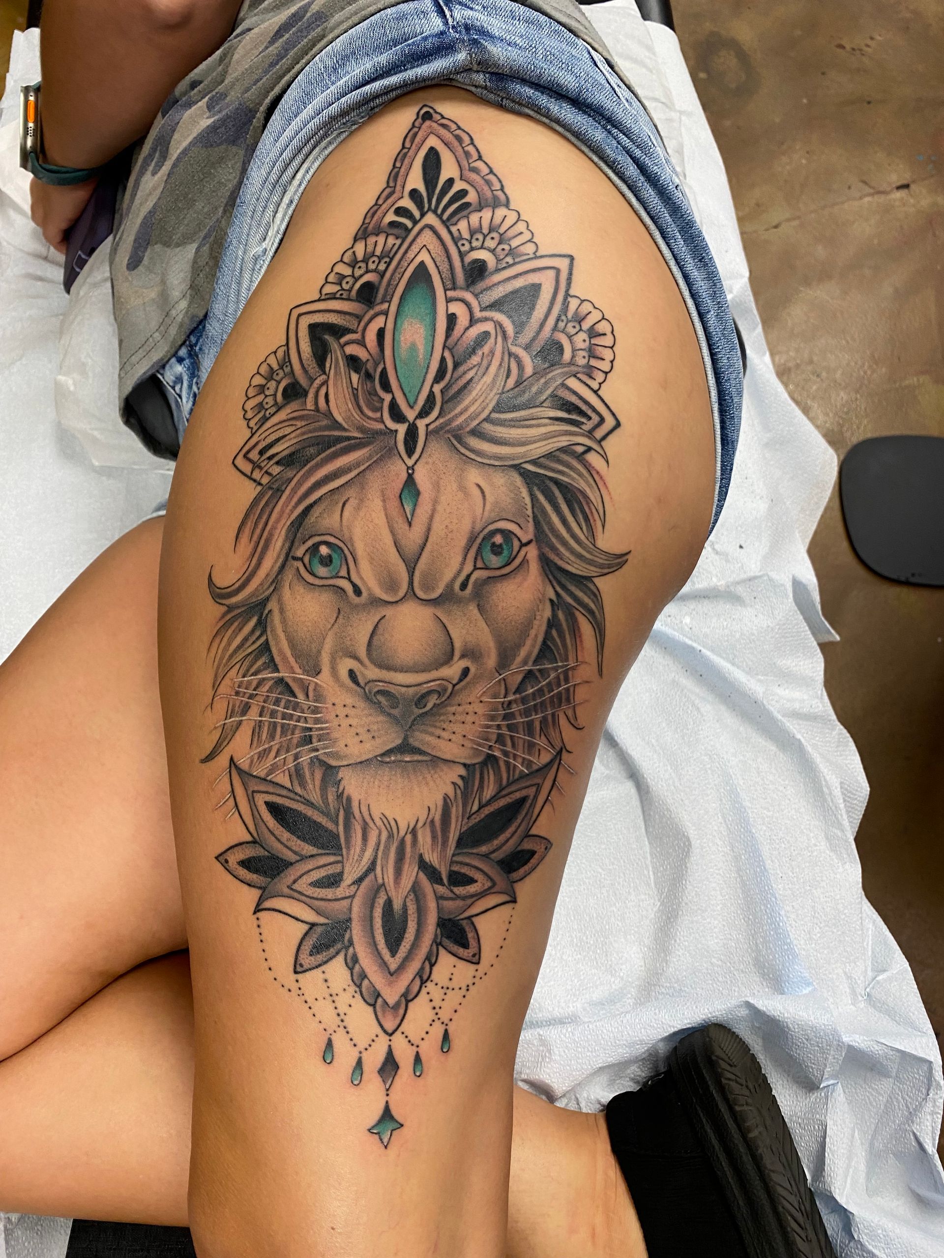 Lion head tattoo with ornate headpiece and lotus flower on a thigh.