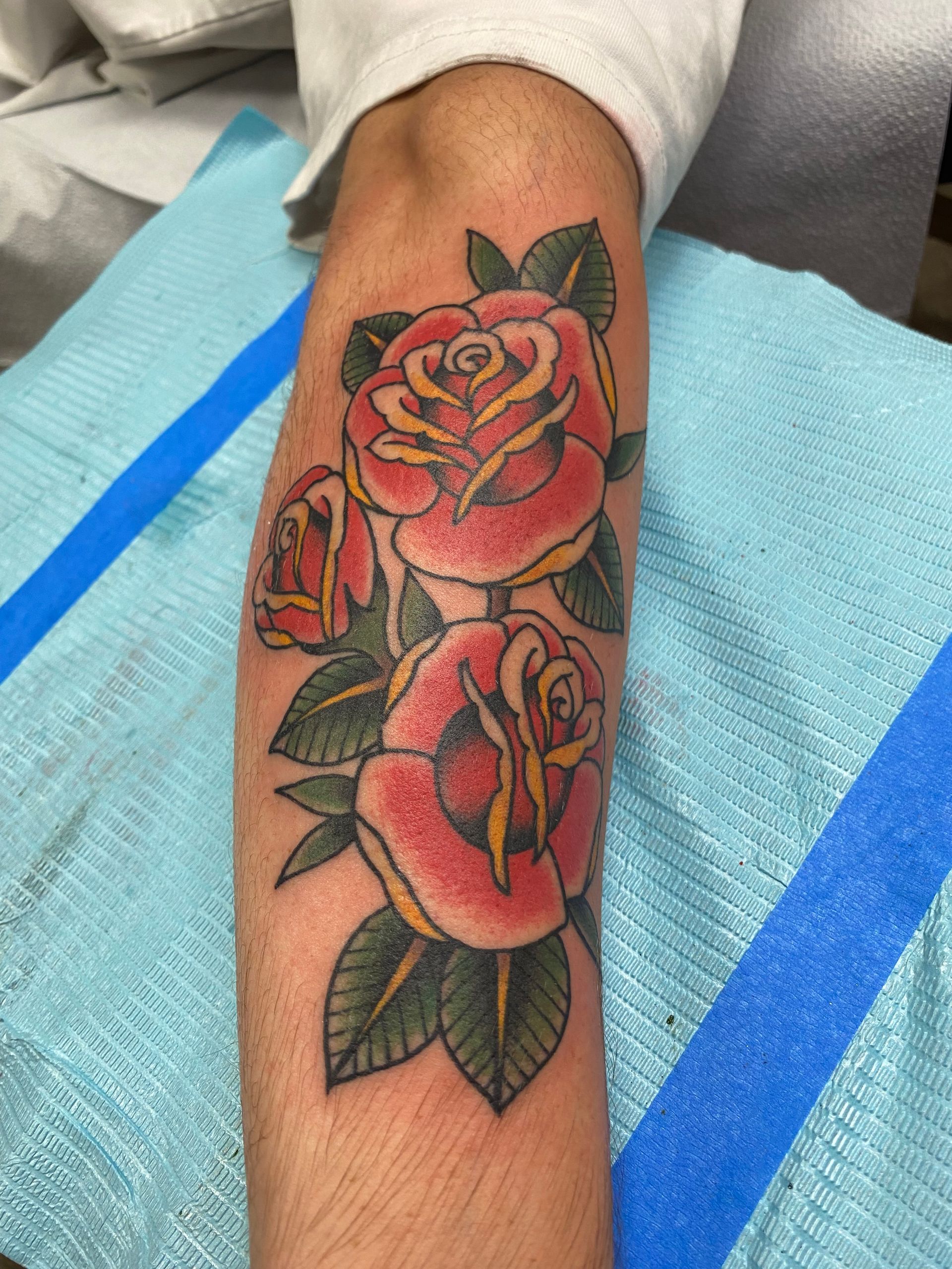 Tattoo of three red roses with yellow centers and green leaves on a forearm, lying on a blue surface.