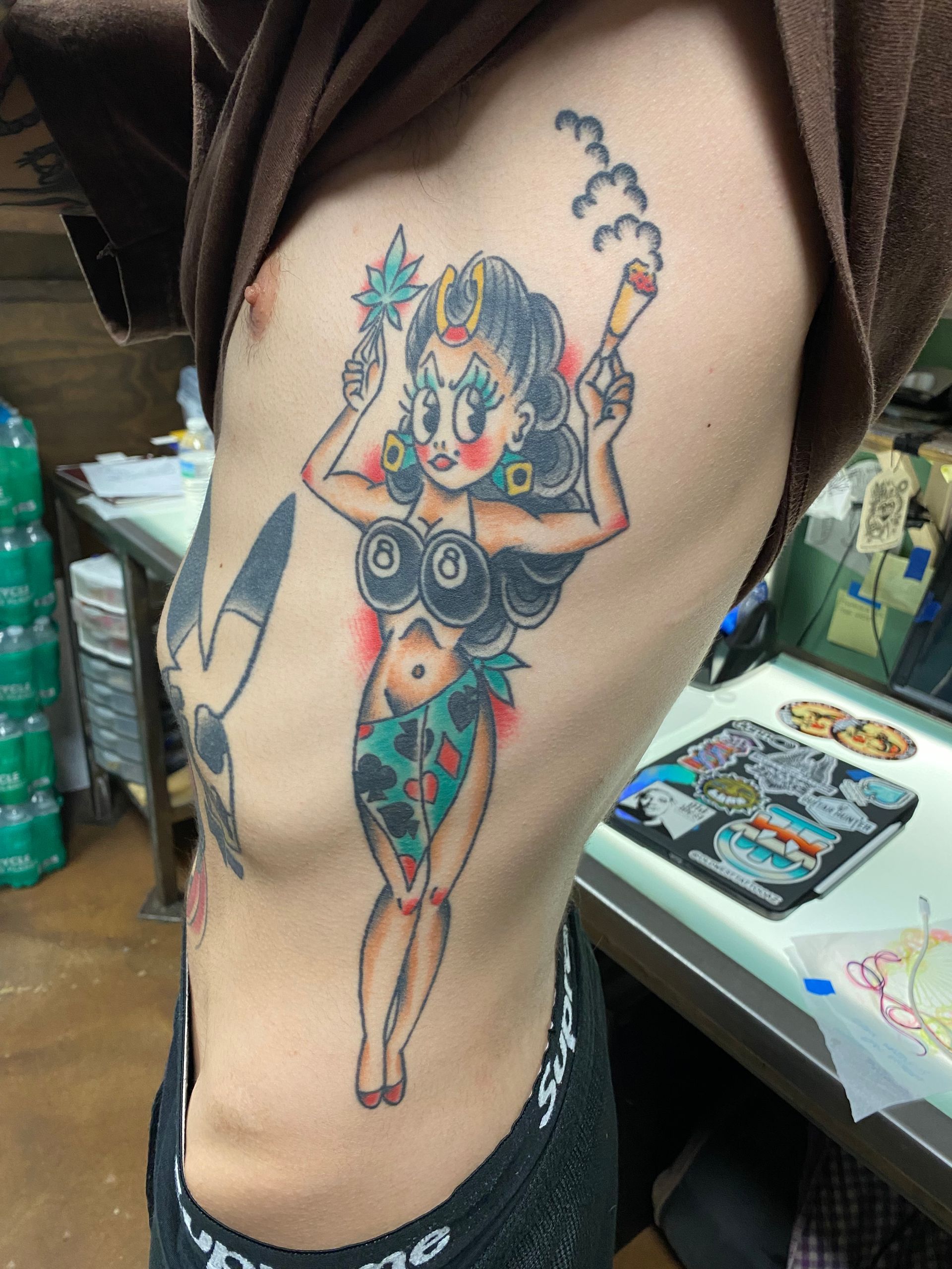 Tattoo on side of torso: woman with cannabis leaf crown holding two lit joints.