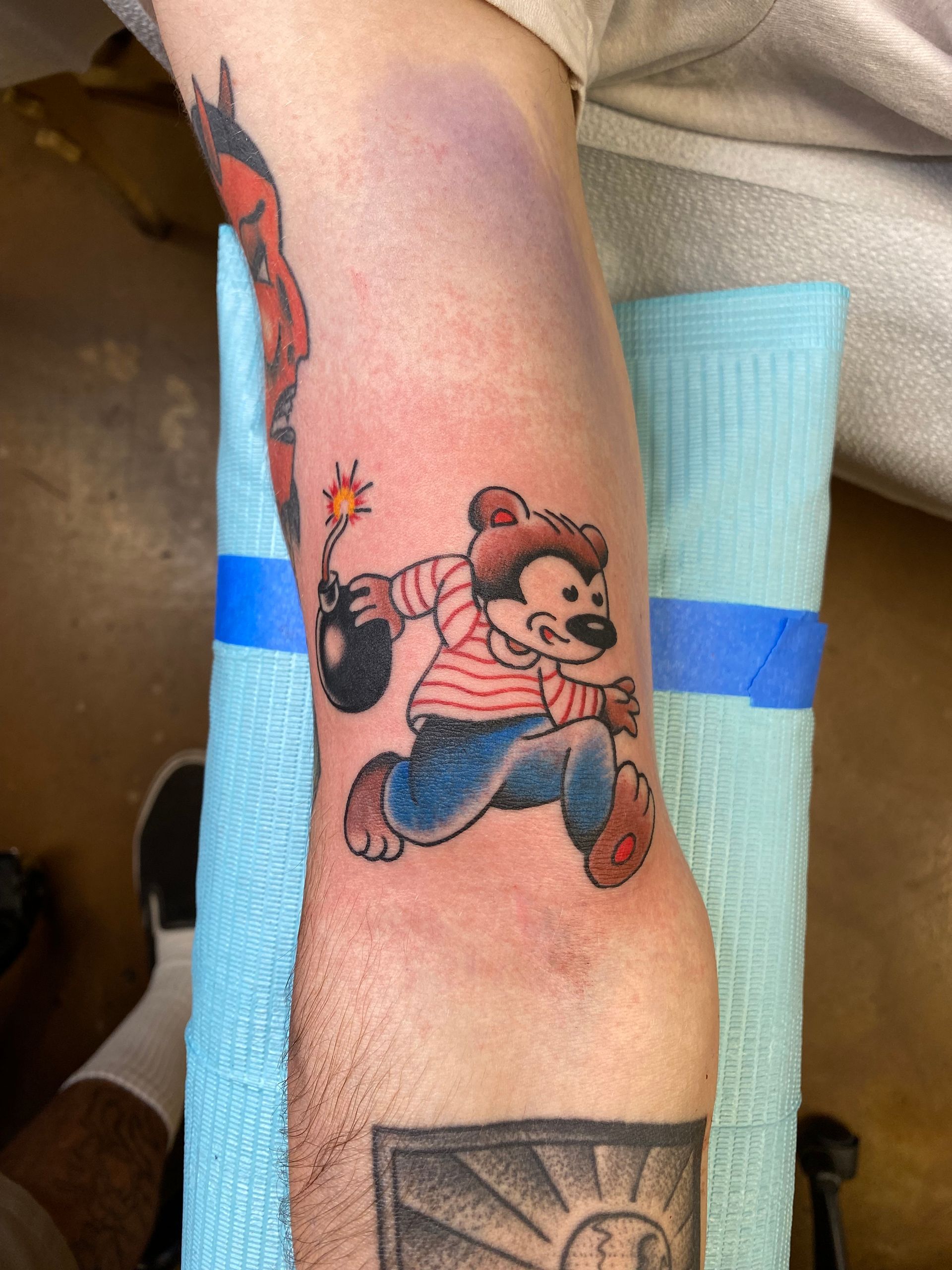 Tattoo of a cartoon bear running with a lit bomb, wearing a striped shirt and jeans, on a person's arm.